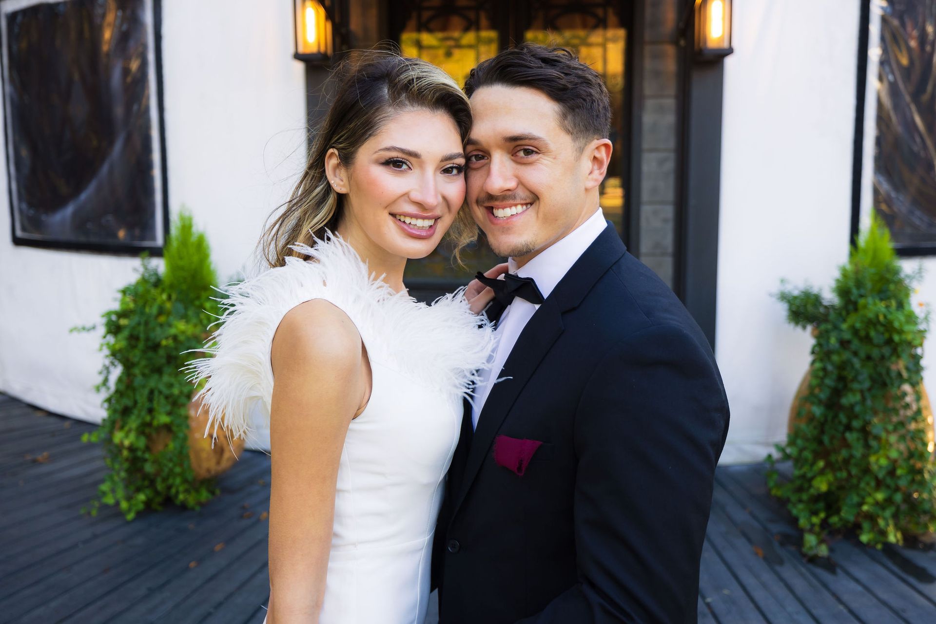 Smiling couple in formal wedding attire standing together outside a building with lanterns and greenery.