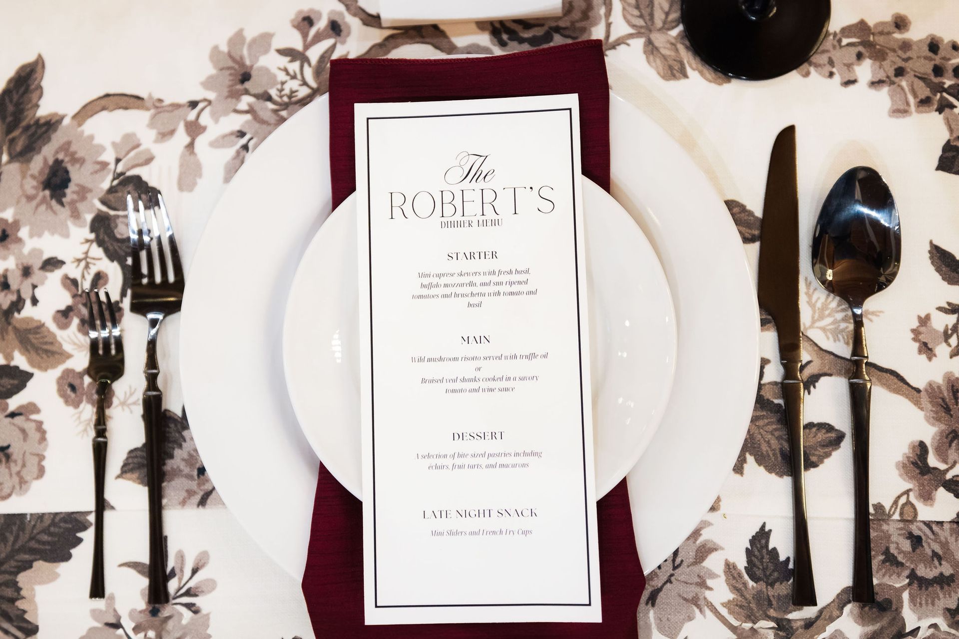 Elegant place setting with white plate, burgundy menu card, and silverware on floral tablecloth
