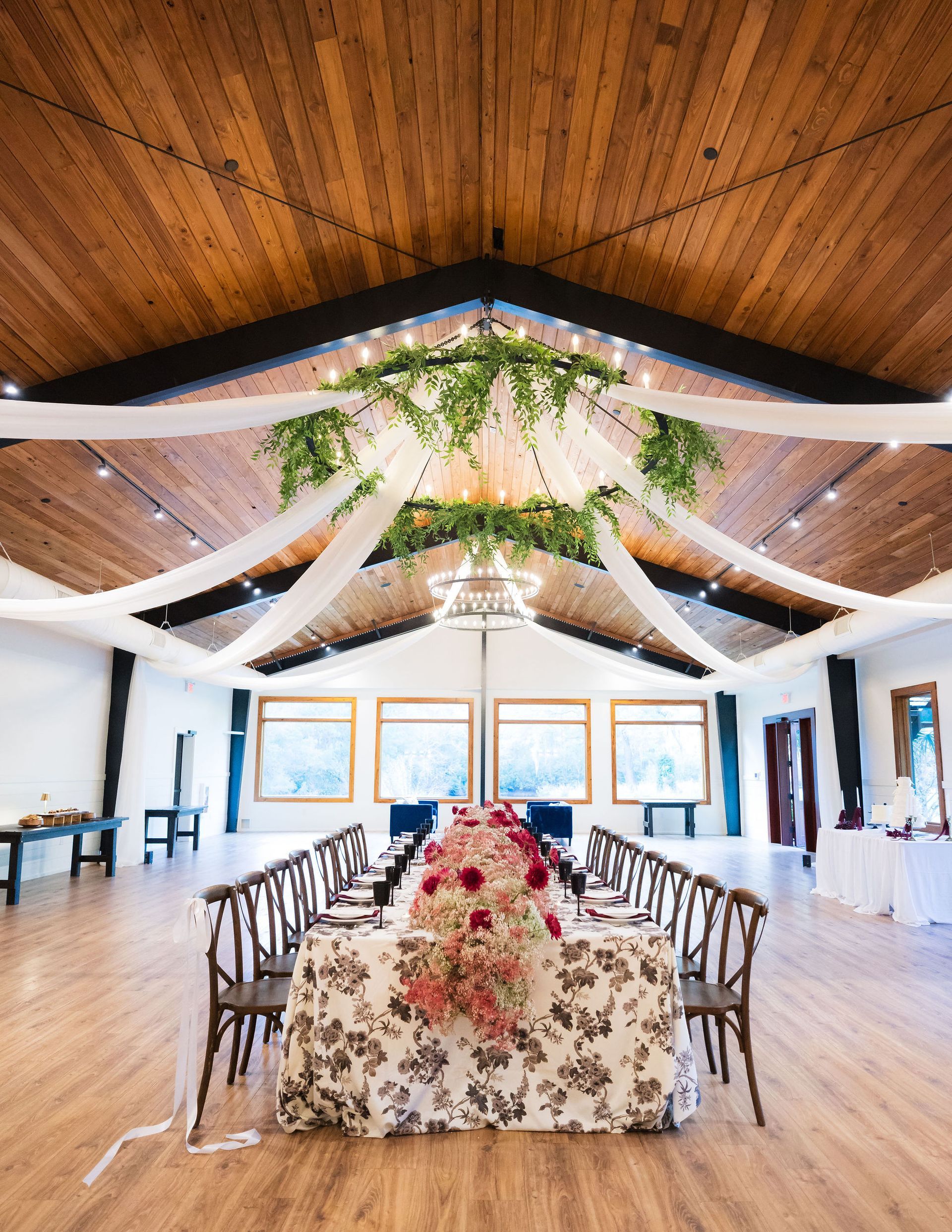 Decorated wedding reception hall with long floral table, white drapes, and greenery under a wood ceiling