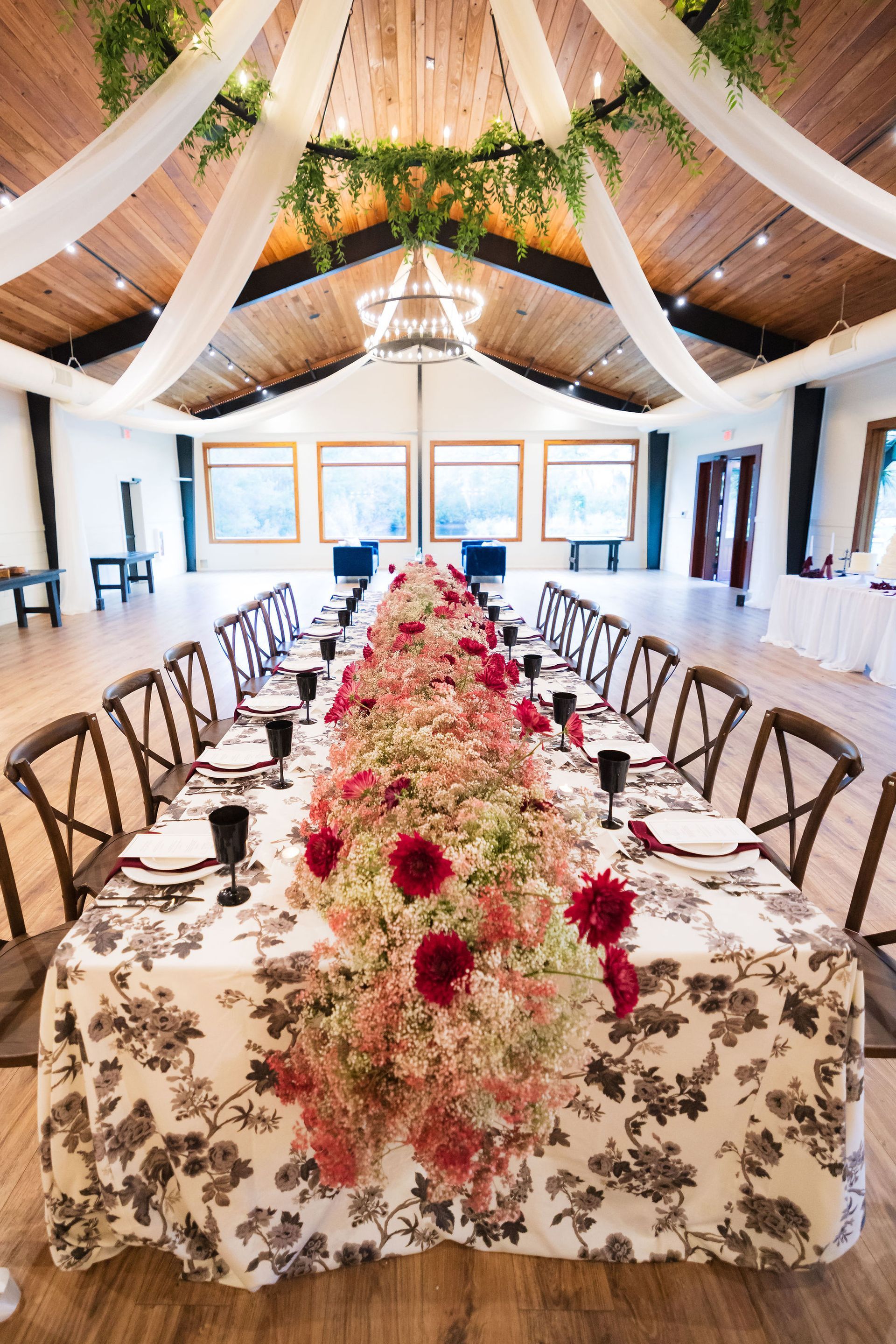 Elegant banquet hall with long floral table, white drapes, and chandeliers under a vaulted wooden ceiling