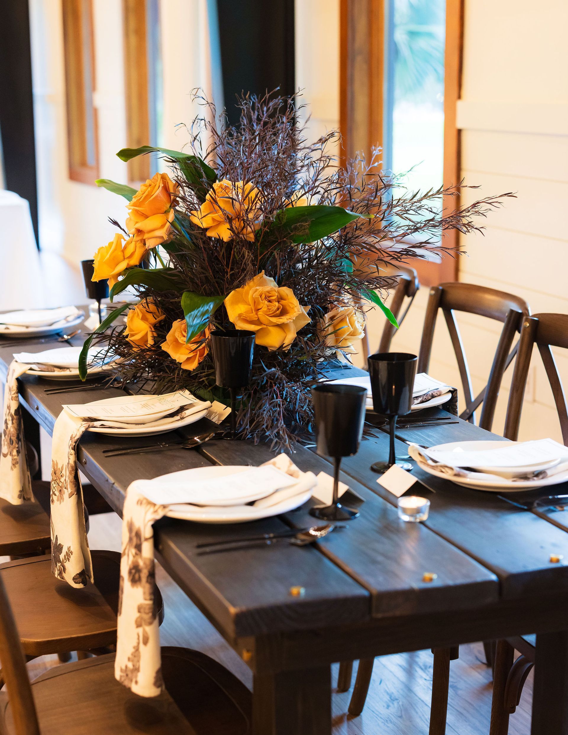Set dining table with black dishes, white napkins, and a yellow-blue floral centerpiece by a window