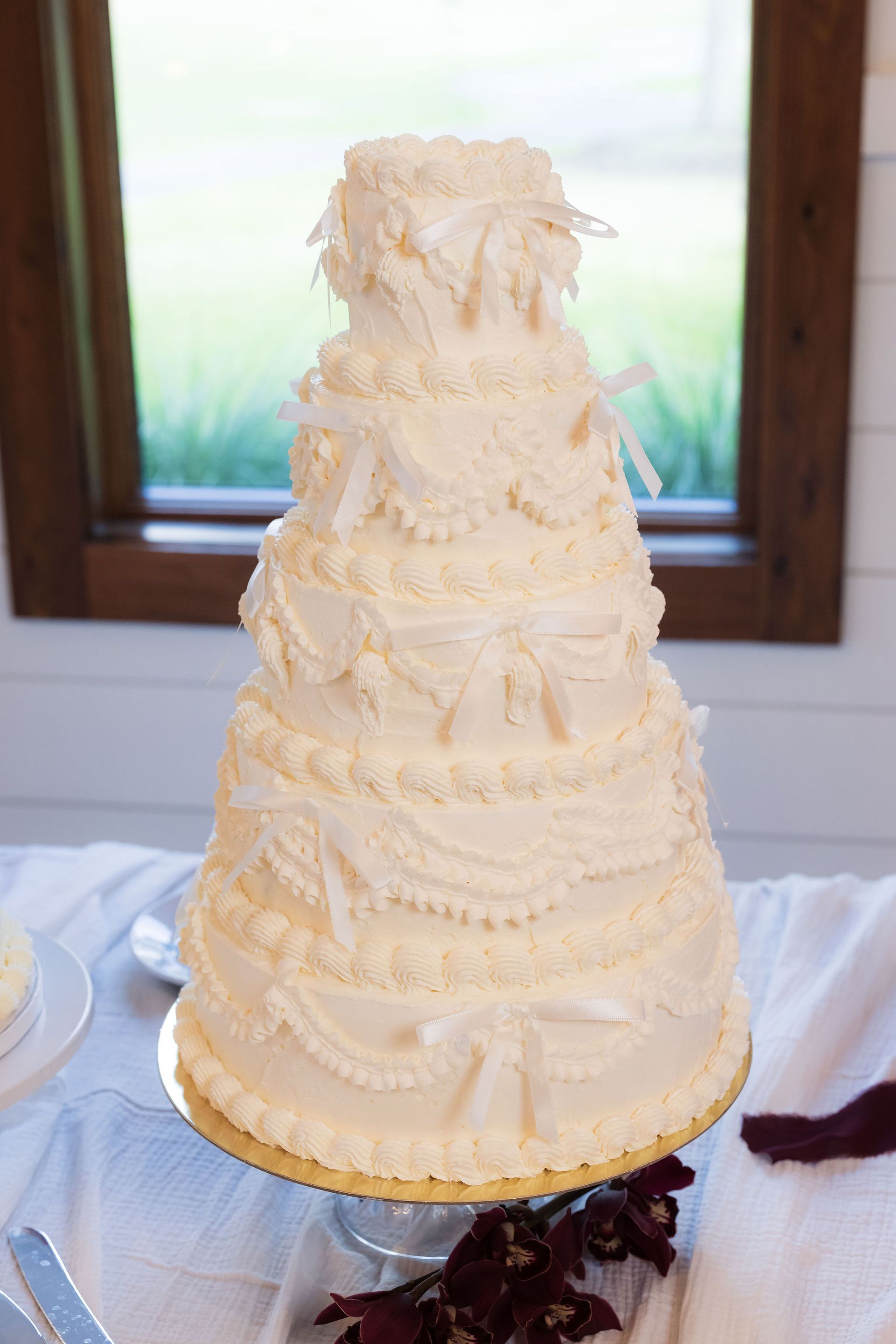 Tall cream-colored wedding cake with lace-like frosting and a ribbon, displayed indoors by a window