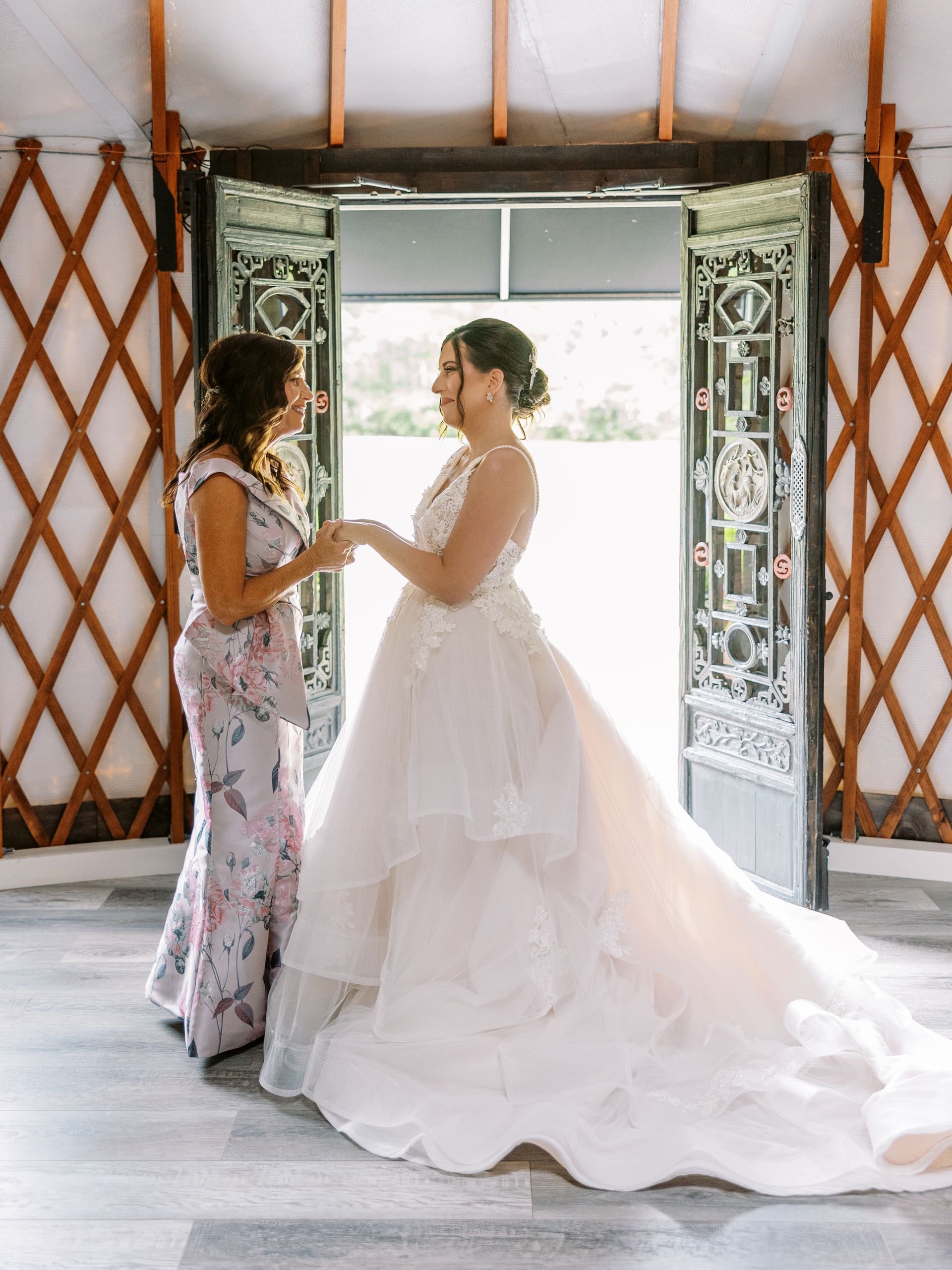 Two women in formal dresses stand holding hands in a bright doorway, one in a white bridal gown.