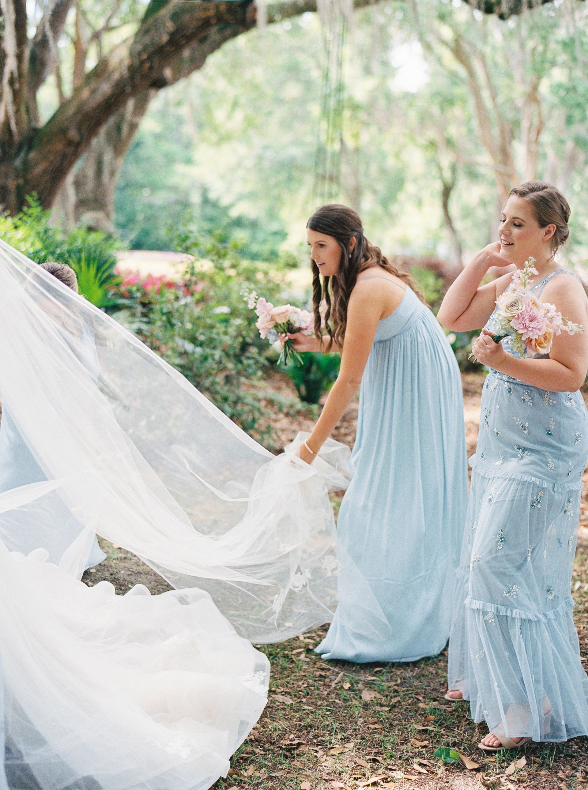 Bridesmaids in pale blue dresses adjusting a bride’s flowing veil in a garden setting