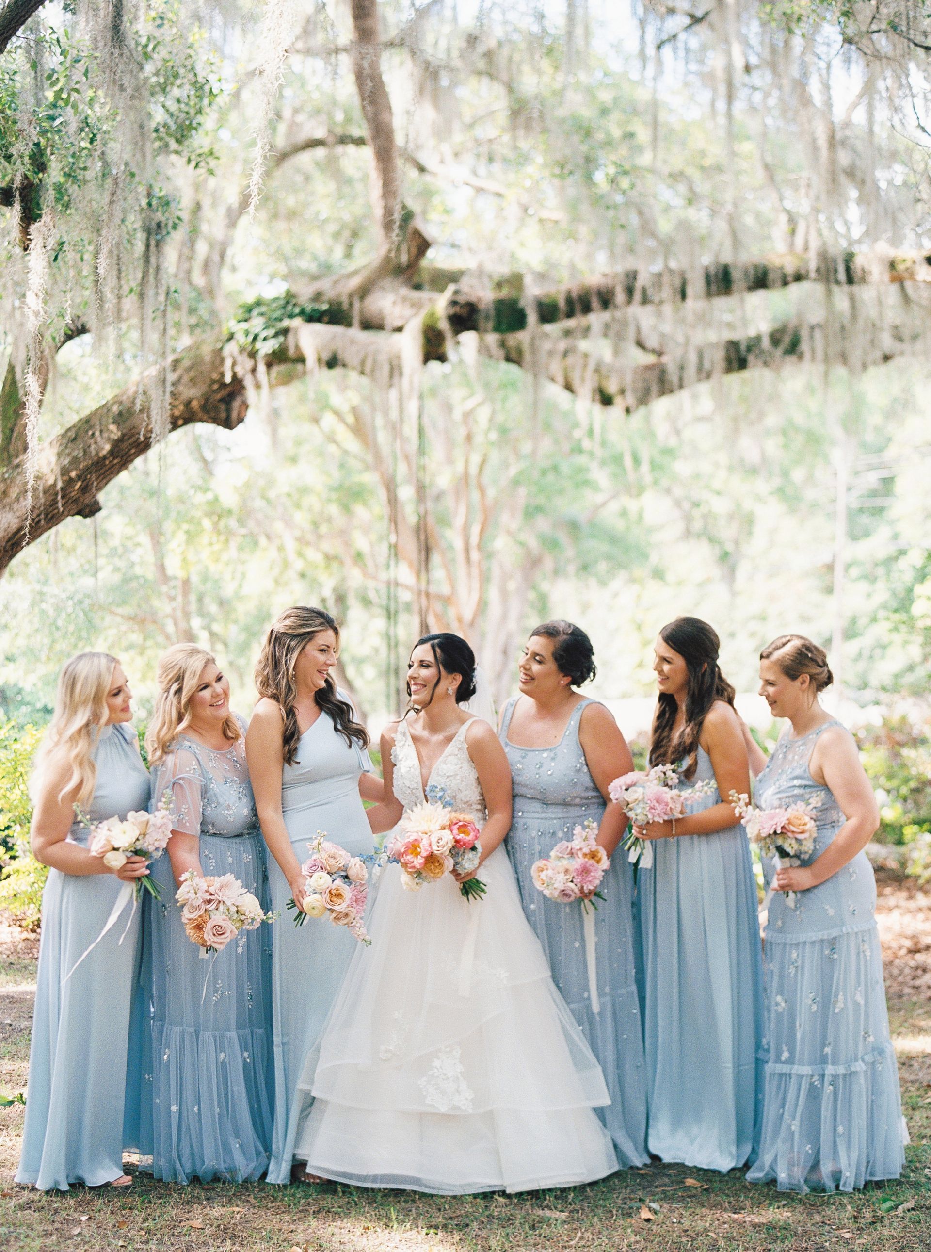 Bride with six bridesmaids in blue dresses holding bouquets outdoors under a tree.
