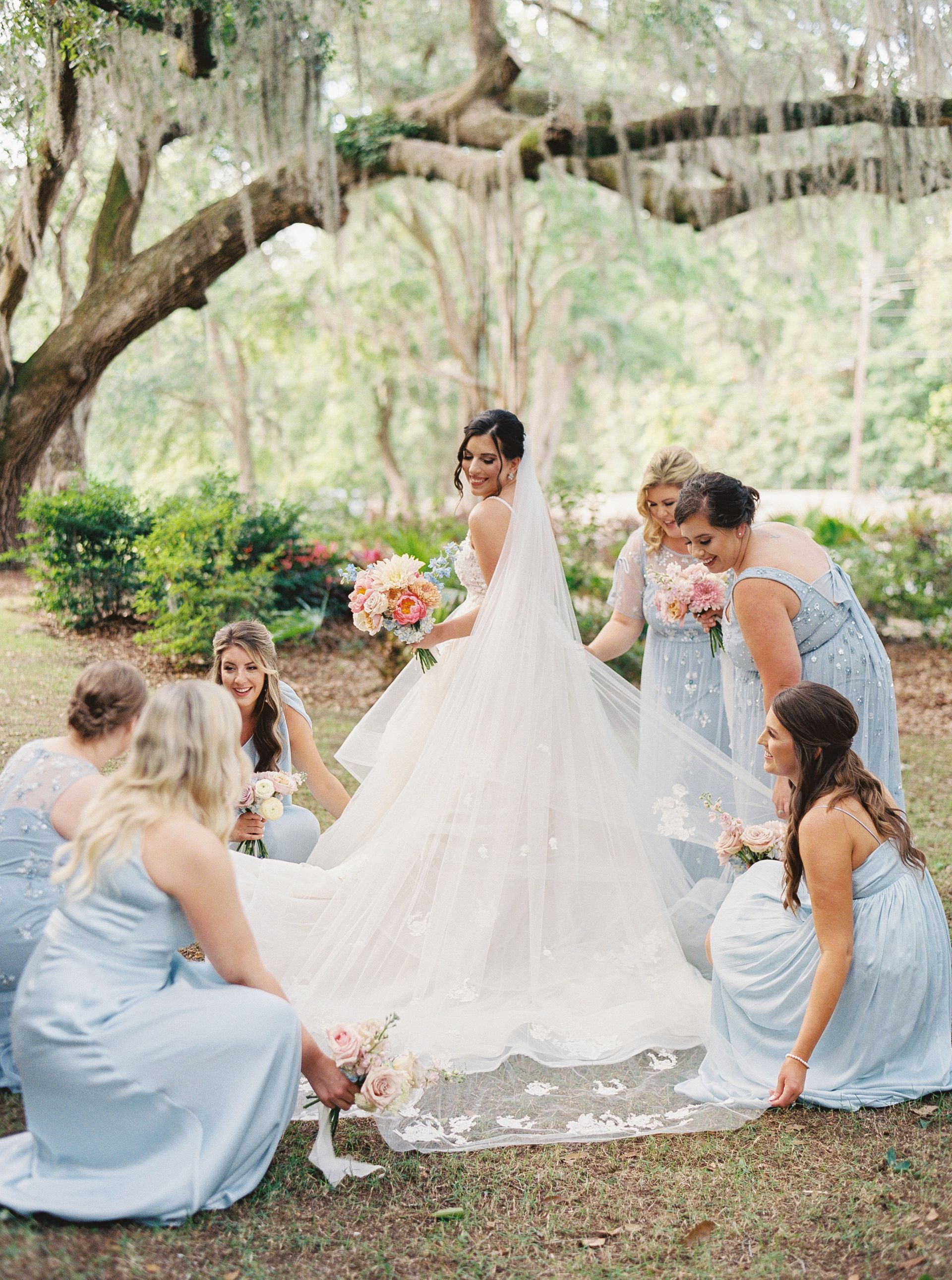 Bride in white gown with bridesmaids in blue dresses under a tree at an outdoor wedding