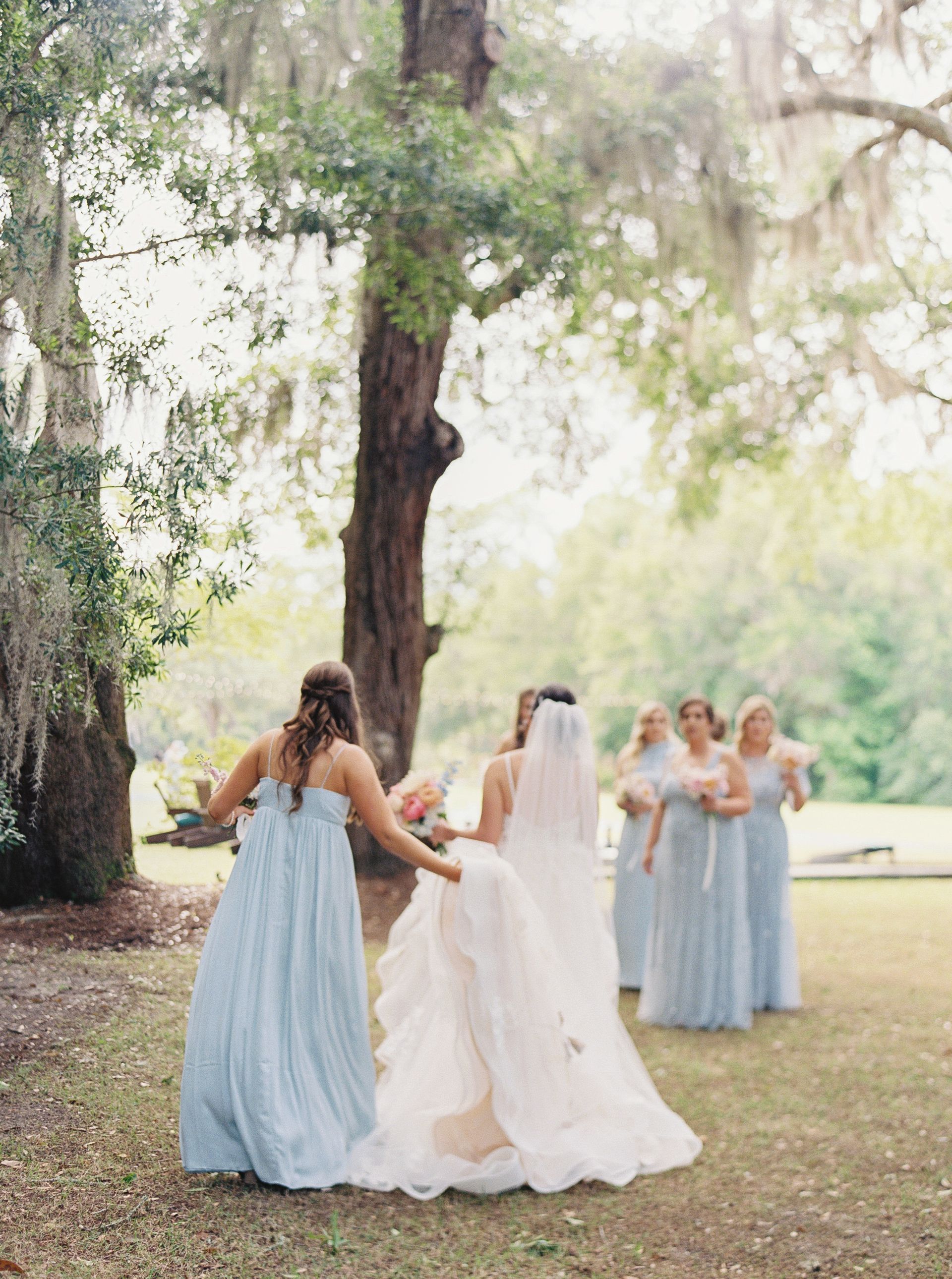 Bridesmaids in light blue dresses surround a bride in a white gown under a large tree outdoors