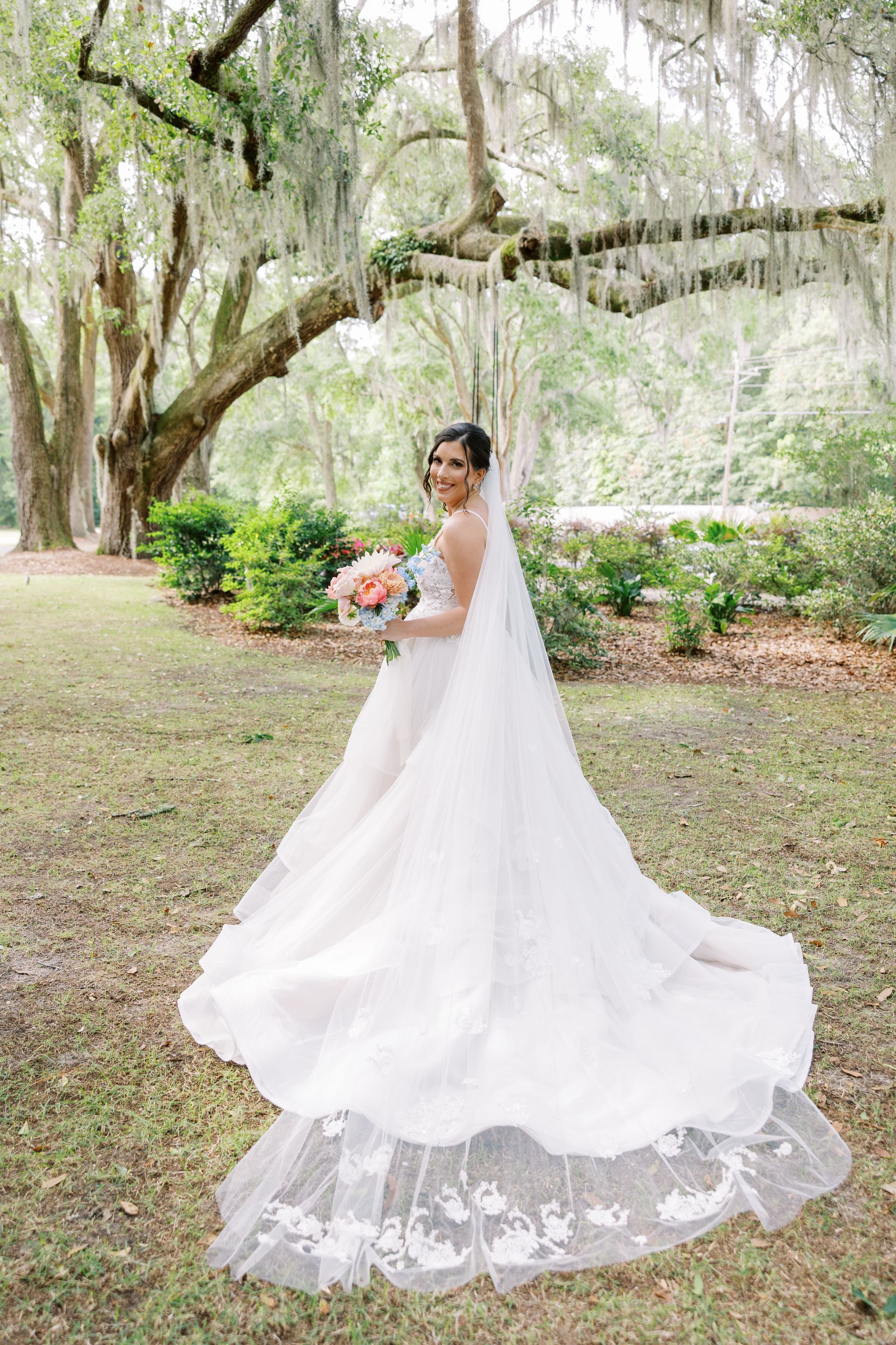 Bride in a white gown holding a bouquet under hanging moss in a garden.