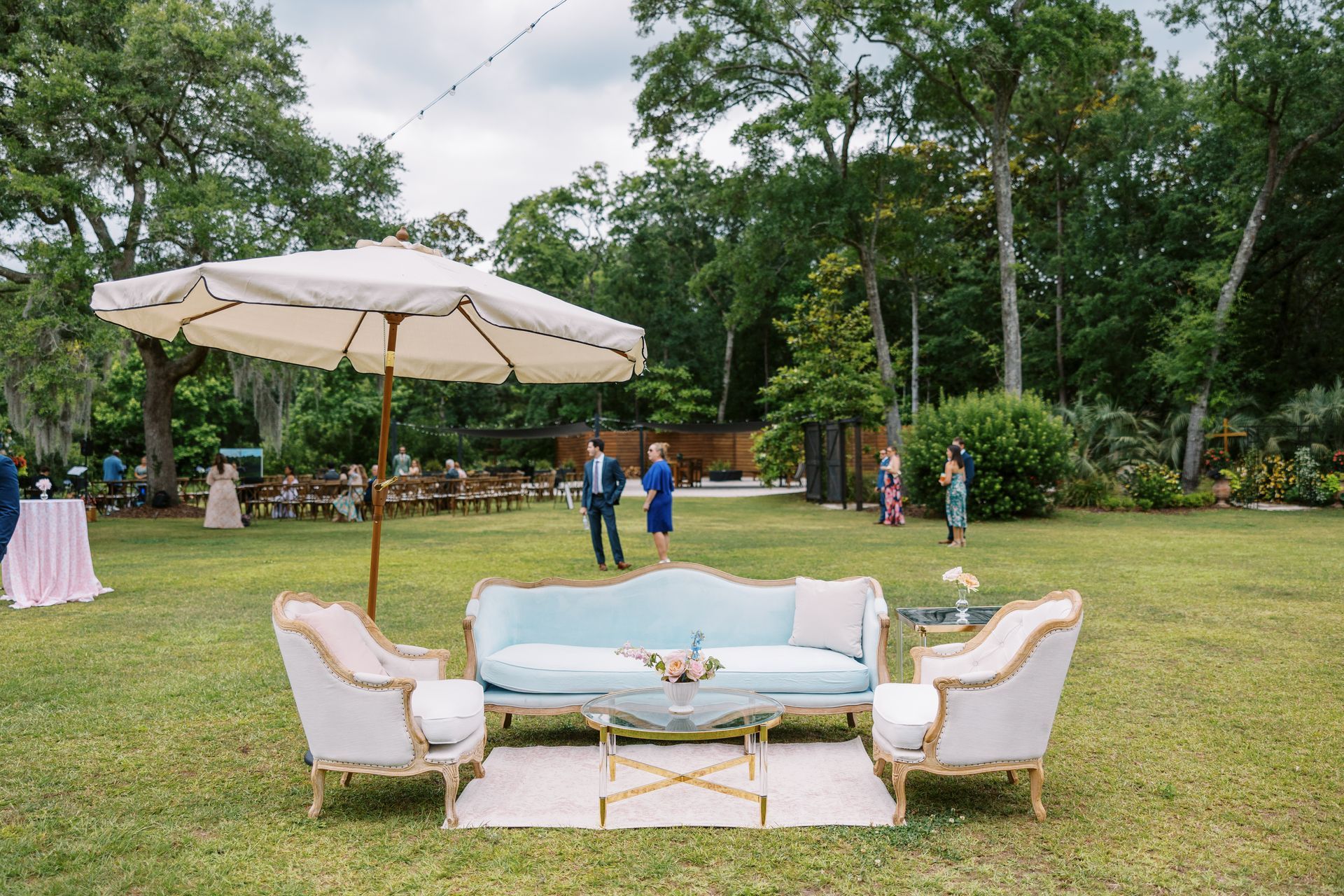Outdoor lounge setup on a grassy lawn with white chairs, a blue sofa, and a large umbrella under string lights.