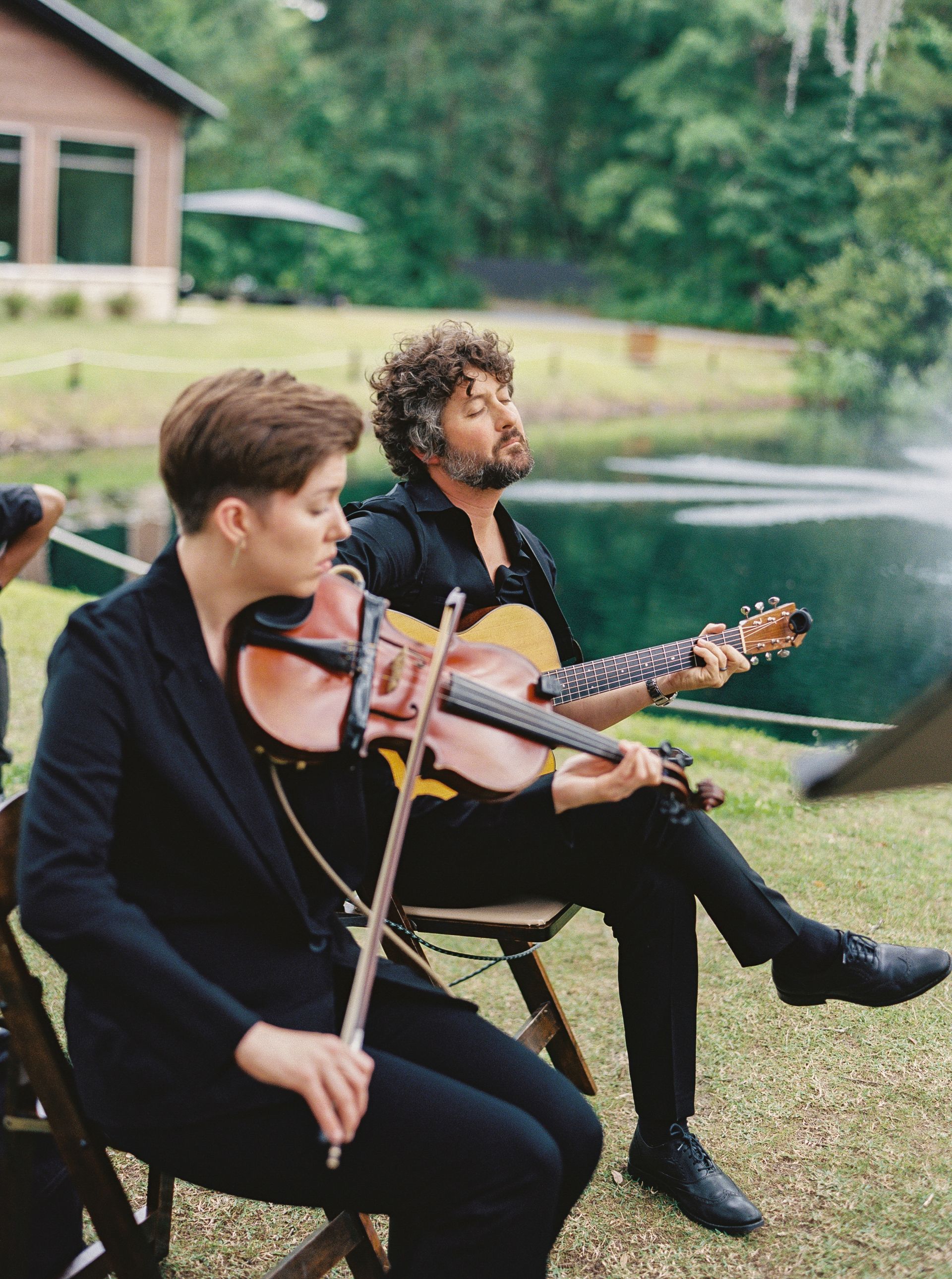 Two musicians in black suits play violin and guitar outdoors beside a lake.