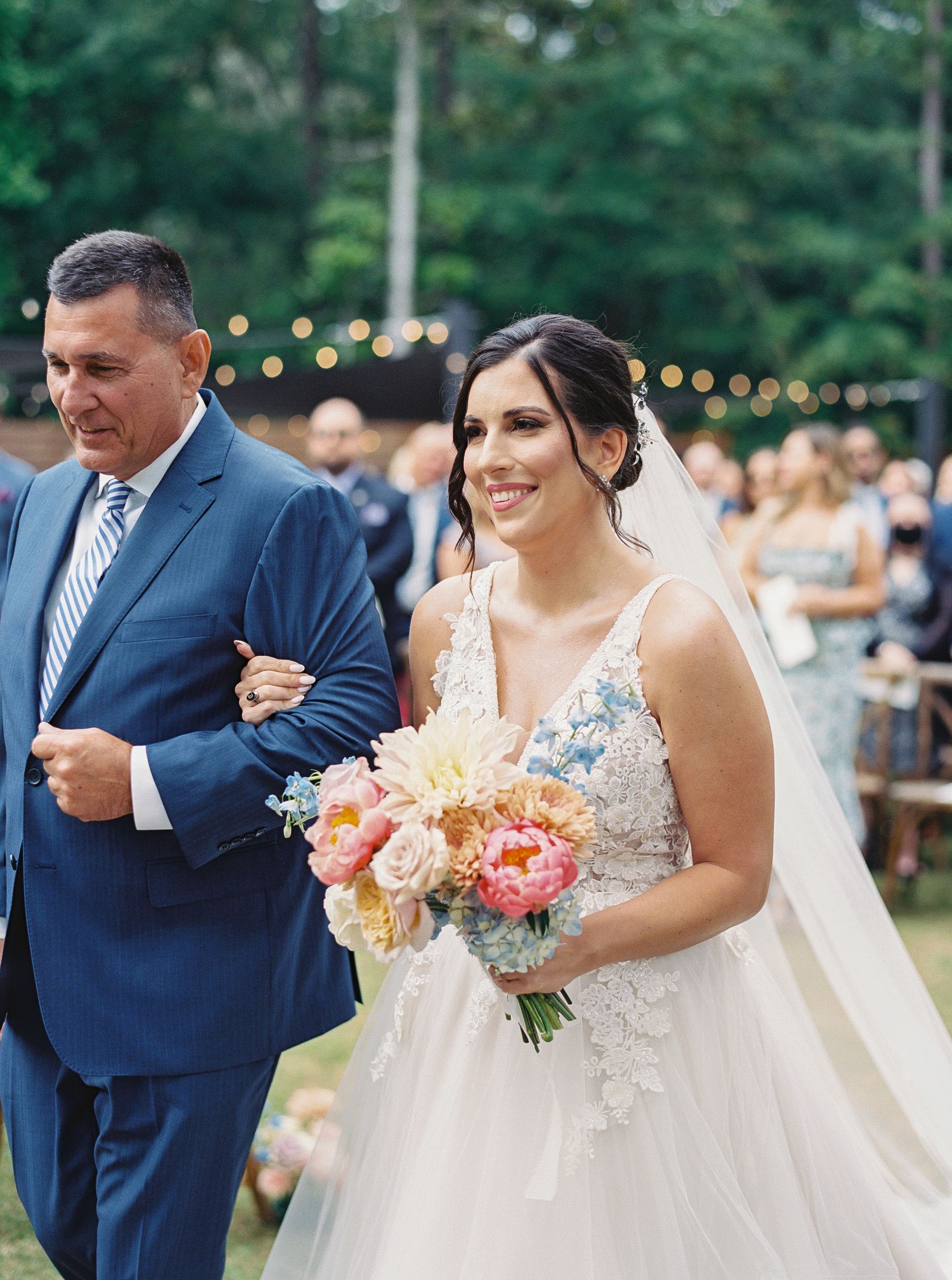Bride in a white lace gown walks arm-in-arm with a man in a blue suit, holding a pink bouquet outdoors.
