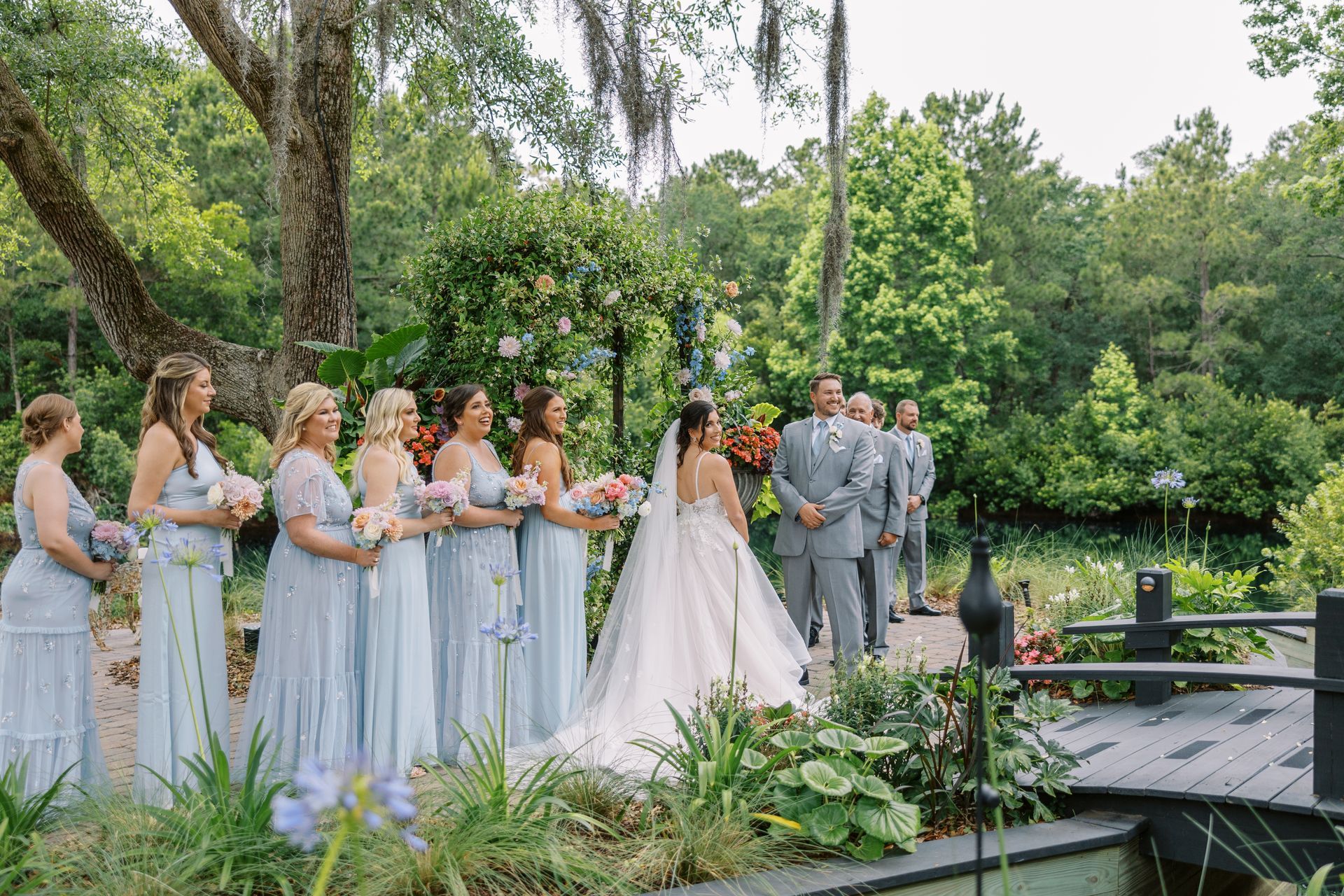 Outdoor wedding ceremony with bridal party in pale blue dresses and gray suits, bride under a floral arch