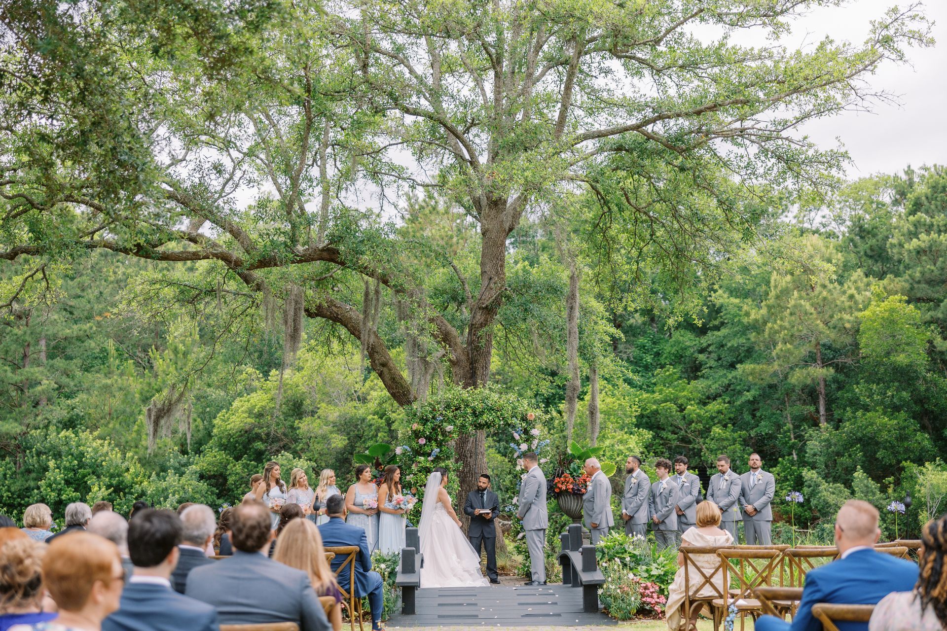 Outdoor wedding ceremony under a large tree, with bride and groom at the altar and guests seated nearby.