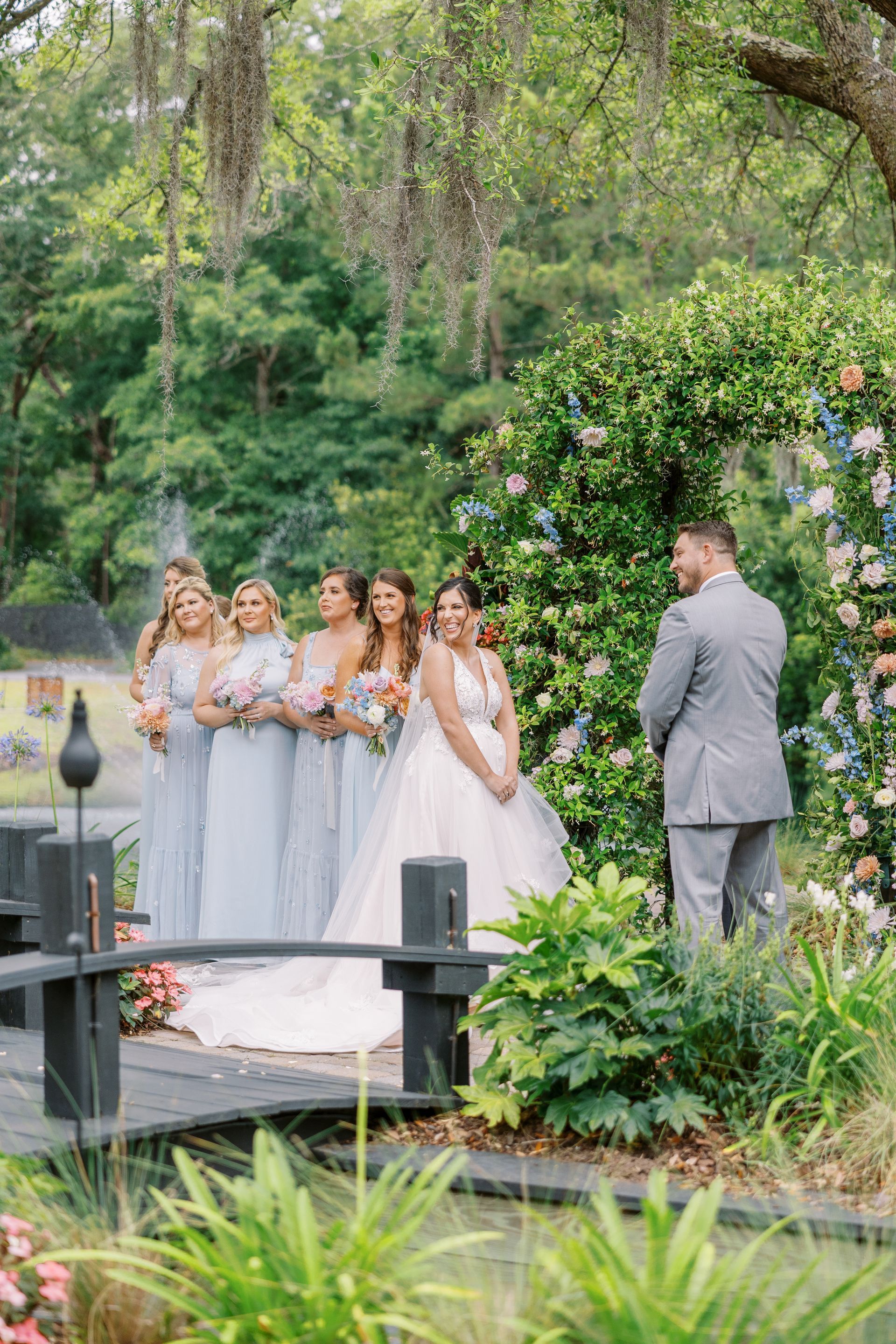 Bridesmaids and bride at an outdoor wedding ceremony, with groom waiting nearby in a garden.
