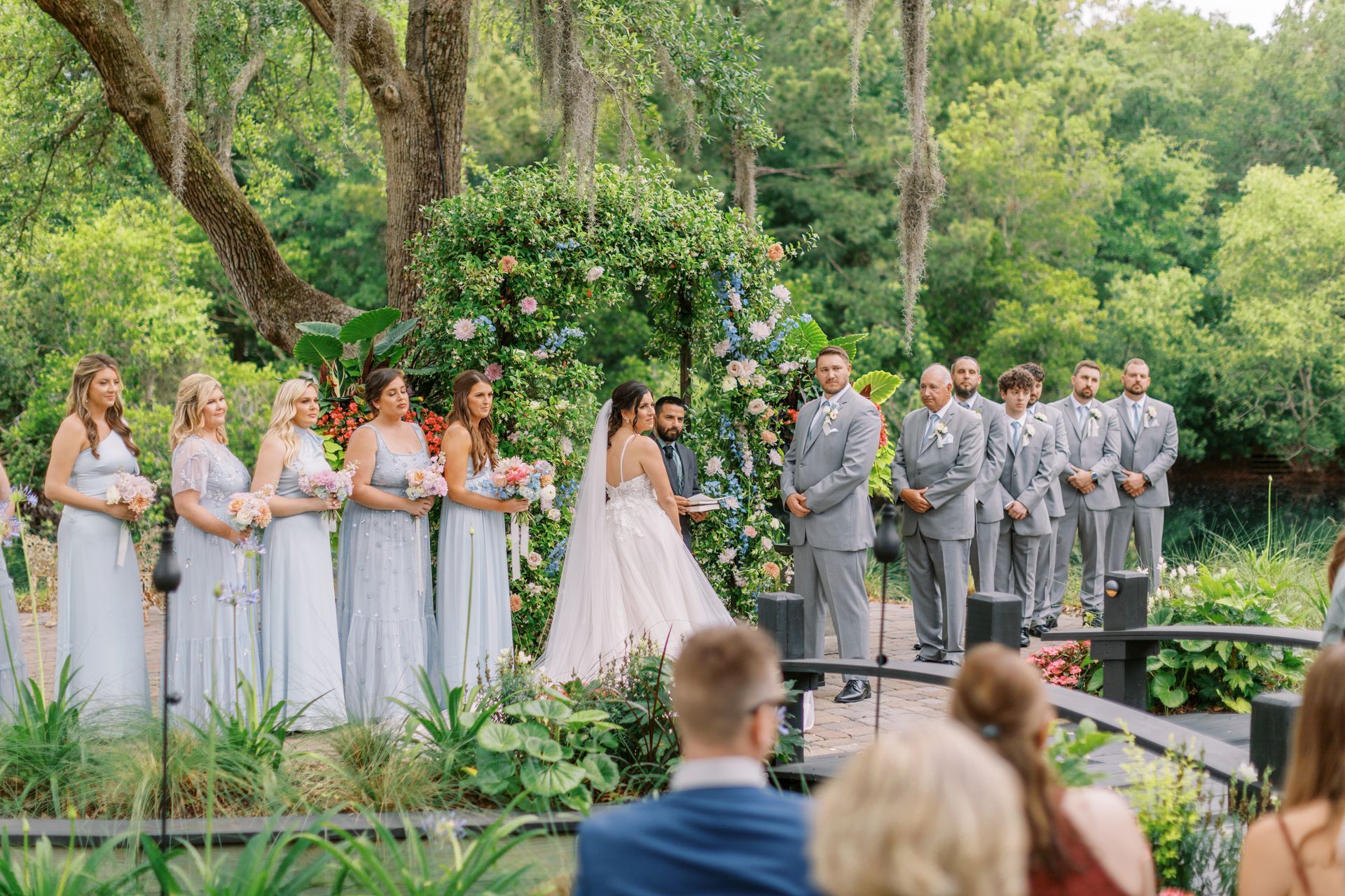 Outdoor wedding ceremony with bride, bridesmaids, and groomsmen in gray suits under trees