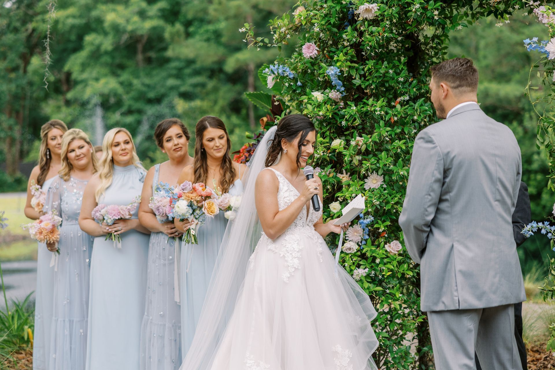 Bride and bridesmaids in lavender dresses posing outdoors with groom in a gray suit during a wedding ceremony