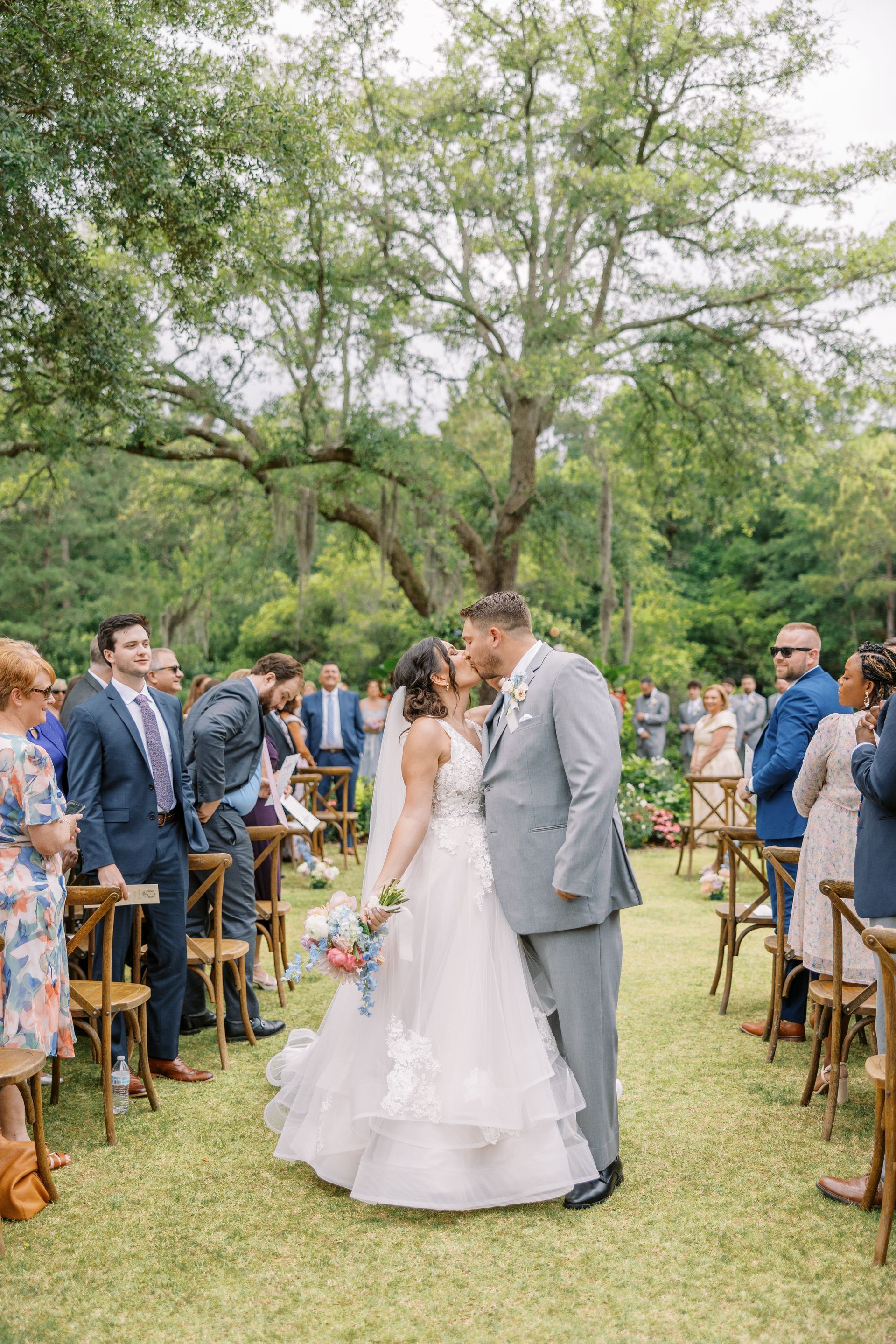 Bride and groom kissing in an outdoor wedding aisle under a large tree, guests standing nearby