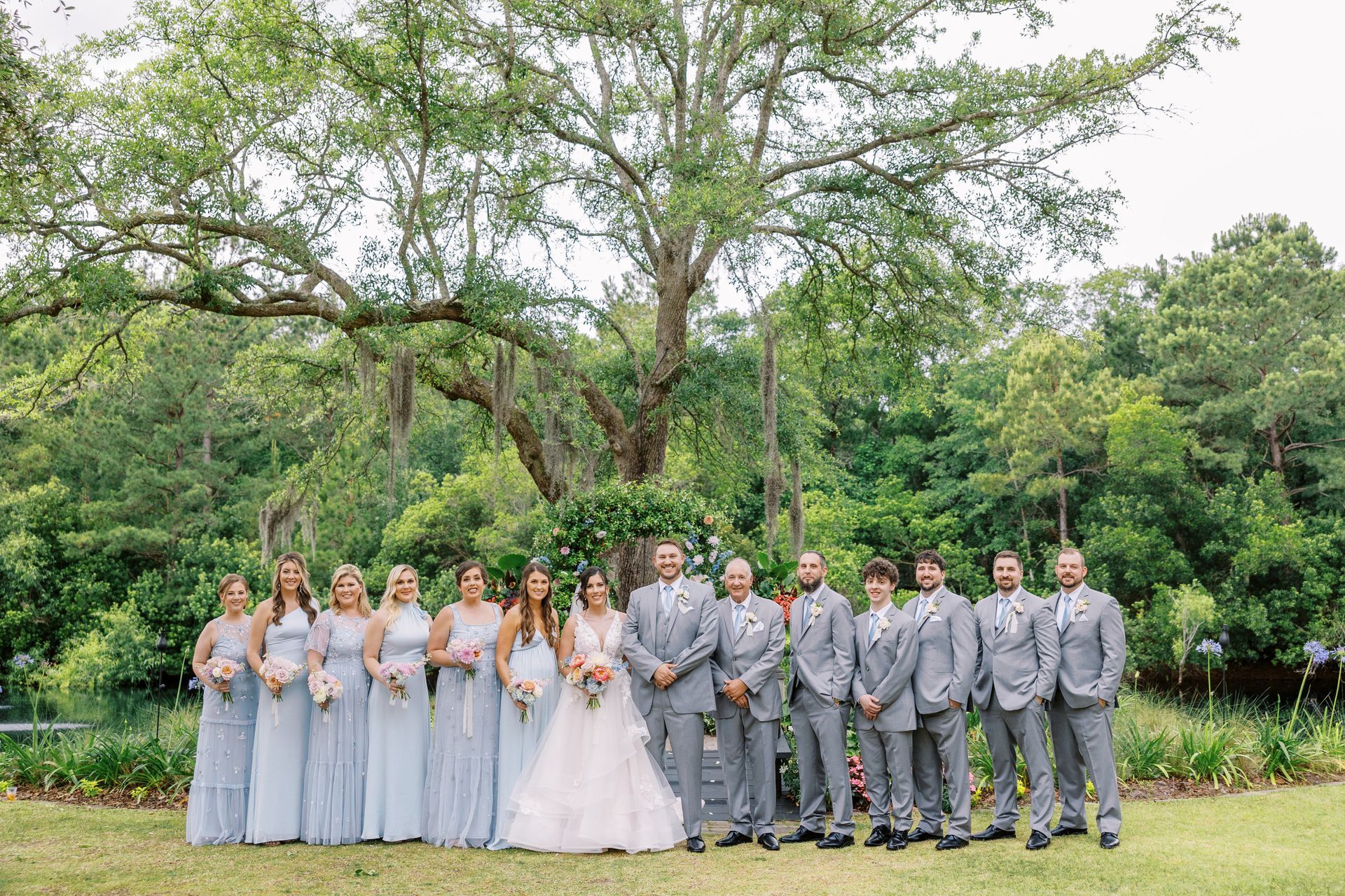 Wedding party posing outdoors under a large tree, bride in white and attendants in gray dresses and suits
