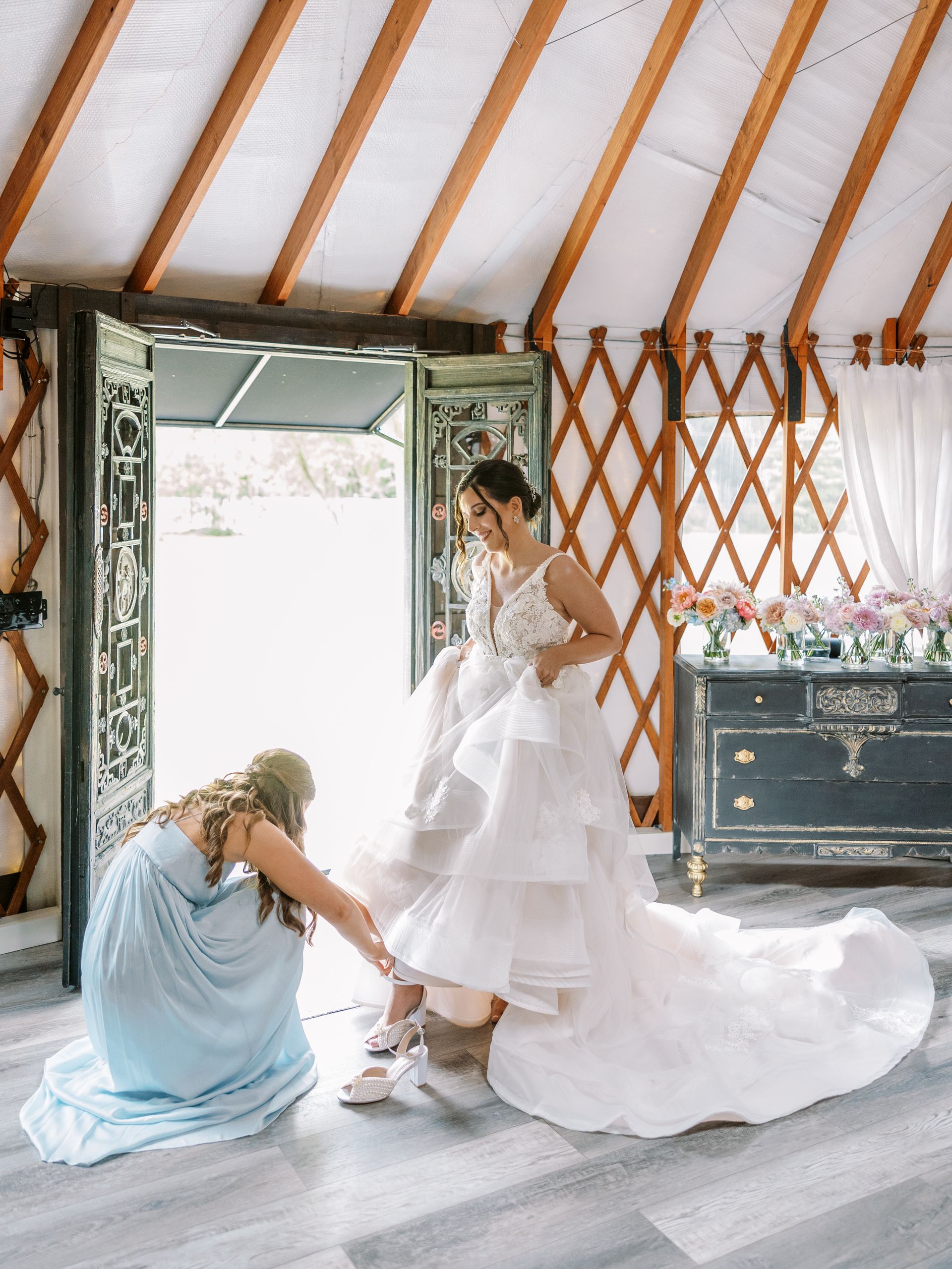 Two people in a rustic room: one adjusts a bride’s white gown, with flowers and an open doorway nearby.