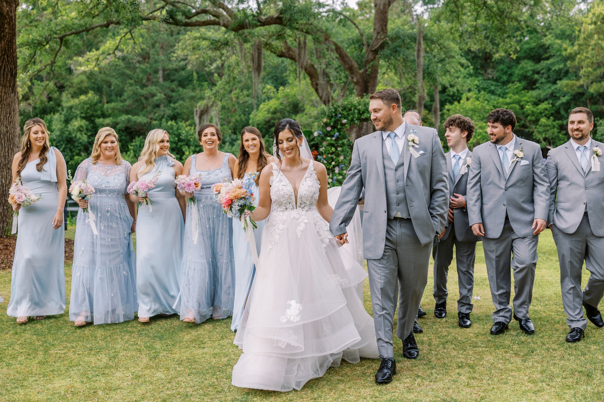 Wedding party walking on grass under trees, bride in white gown with bridesmaids and groomsmen in gray attire