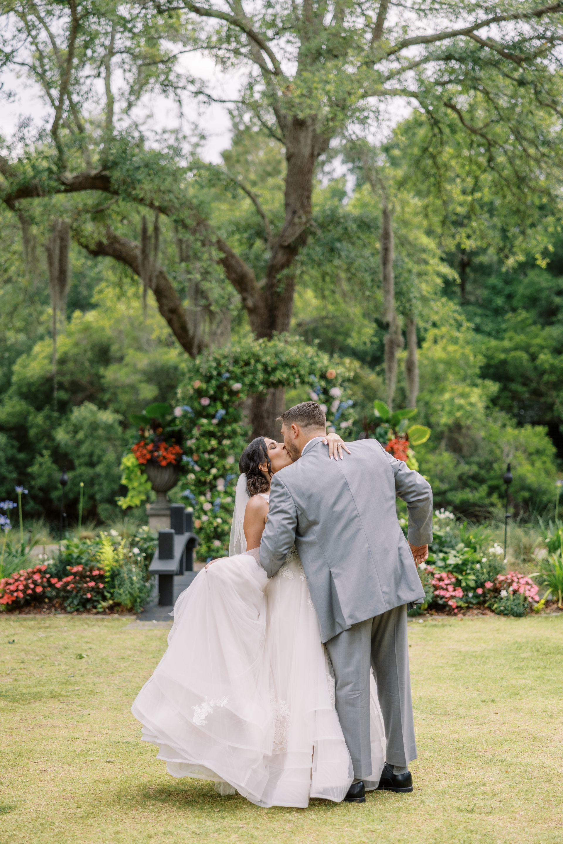 Bride and groom embrace outdoors in a garden surrounded by flowers and tall trees.
