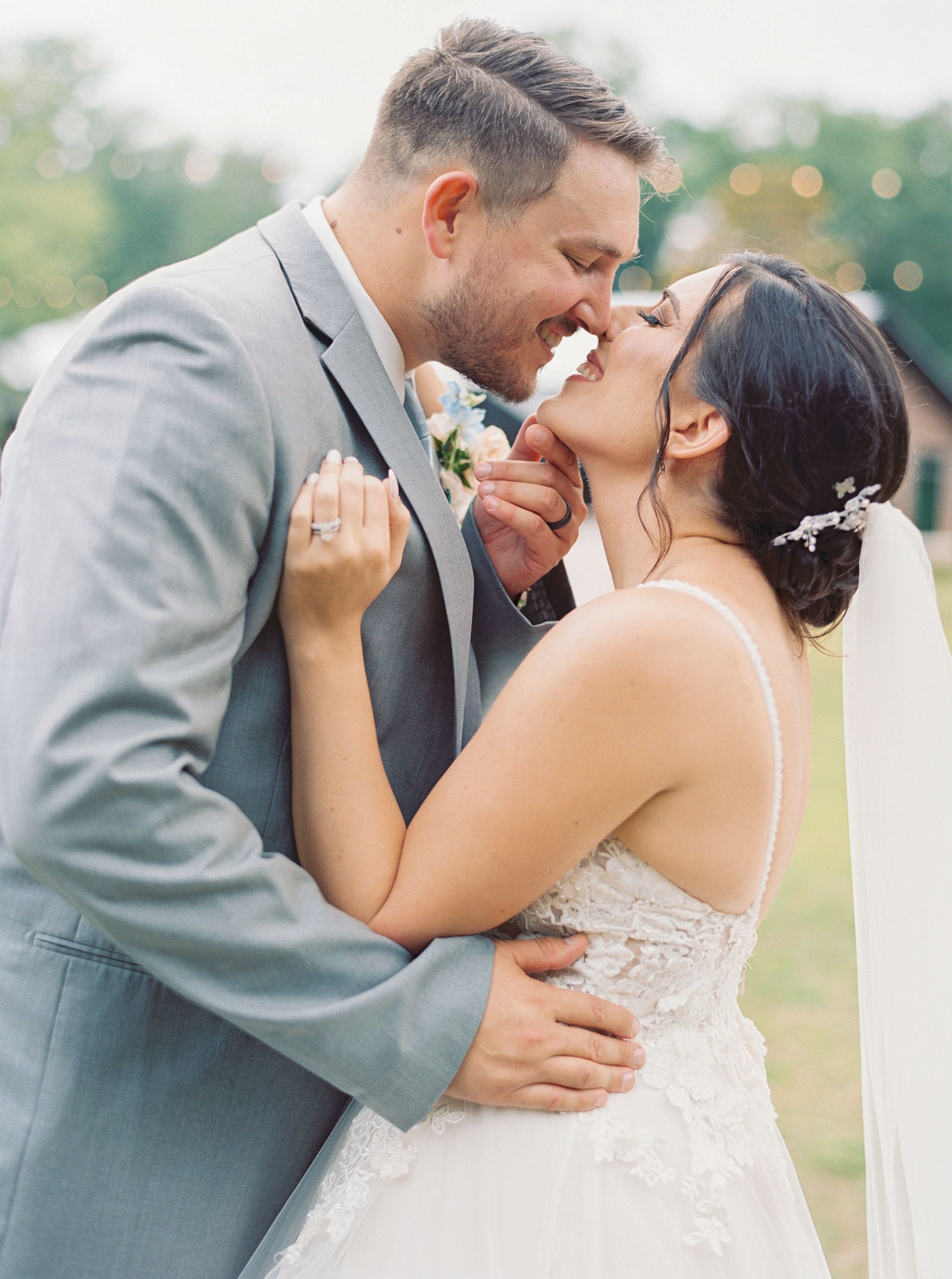 Bride and groom kiss outdoors, with the groom in a light gray suit and the bride in a white gown.