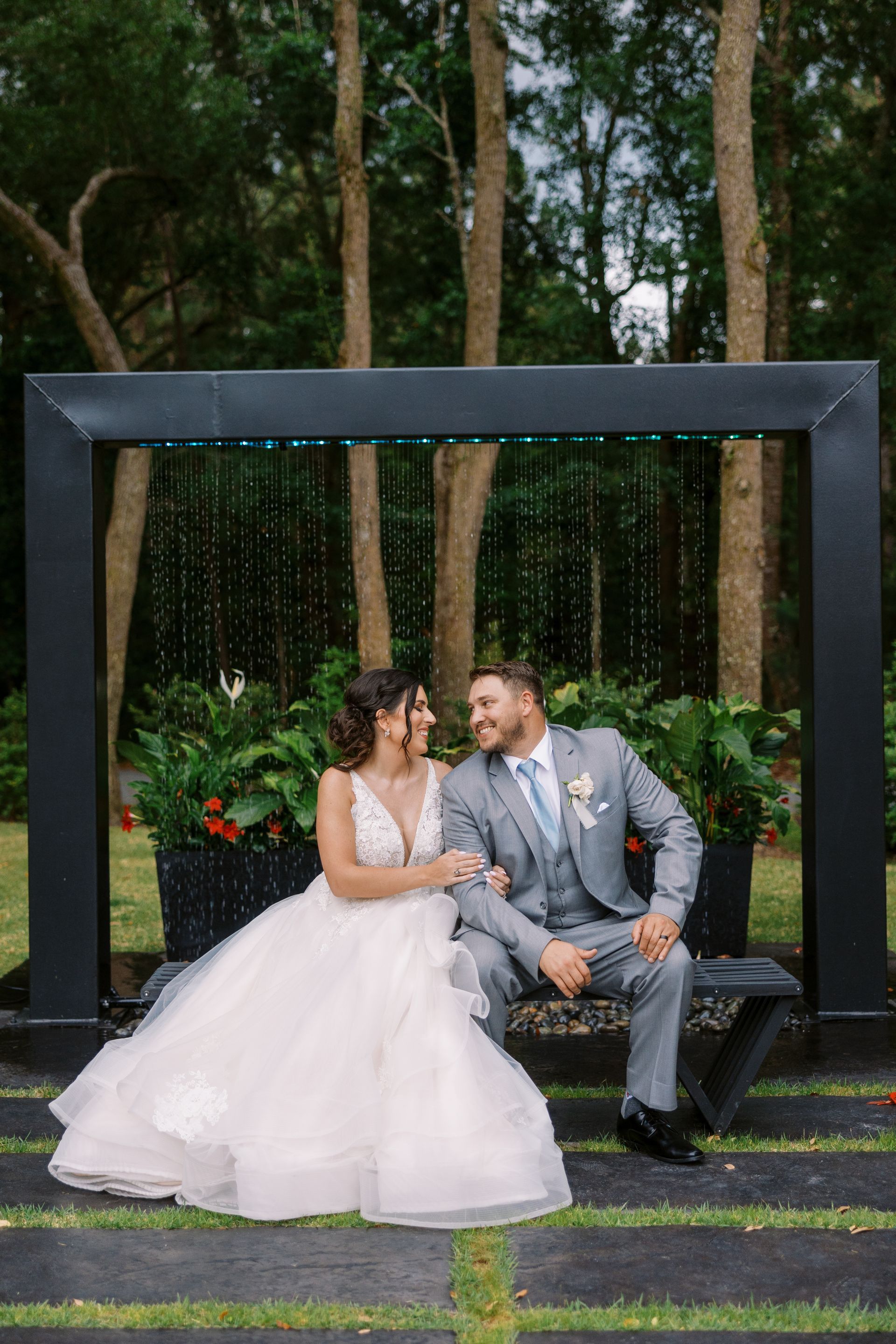 Bride in white gown and groom in gray suit seated on a black bench in a garden, smiling at each other