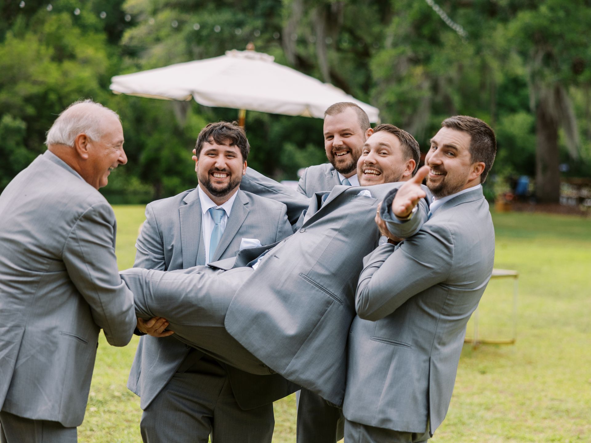 Five men in gray suits laughing outdoors while carrying a man in a suit horizontally