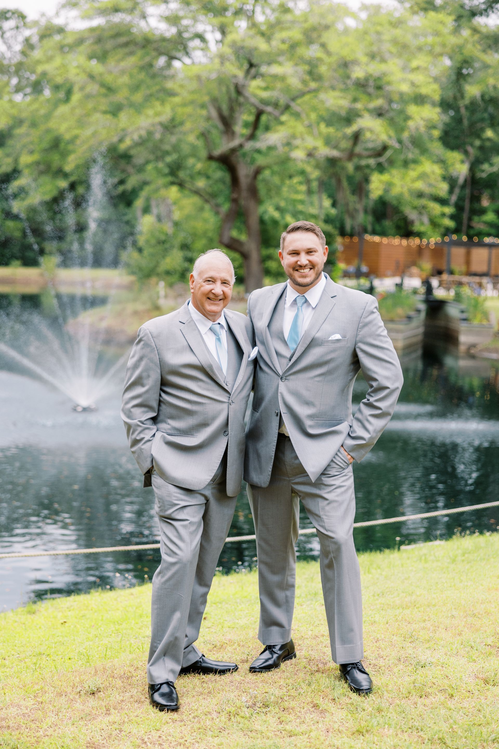 Two men in gray suits standing by a pond with a fountain and trees in the background