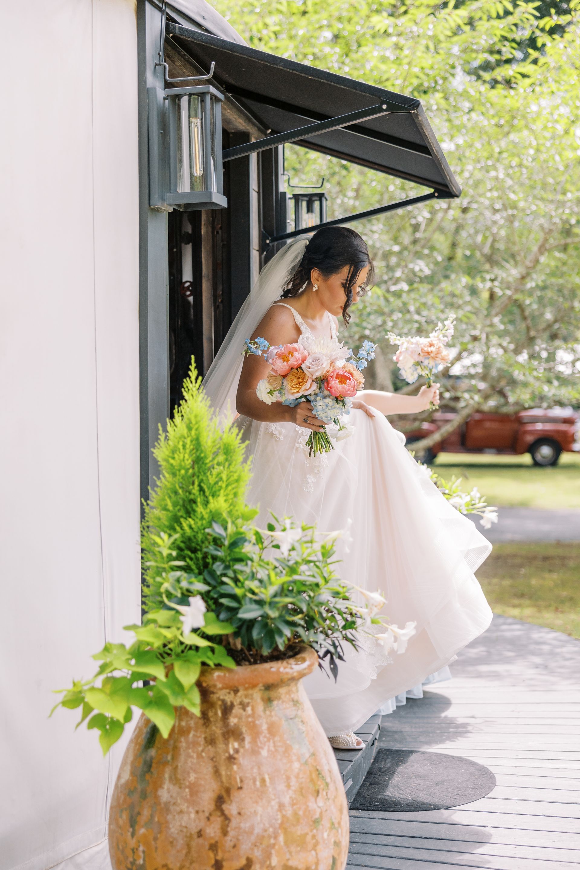 Bride in white gown holding a bouquet, stepping out onto a porch beside potted greenery.