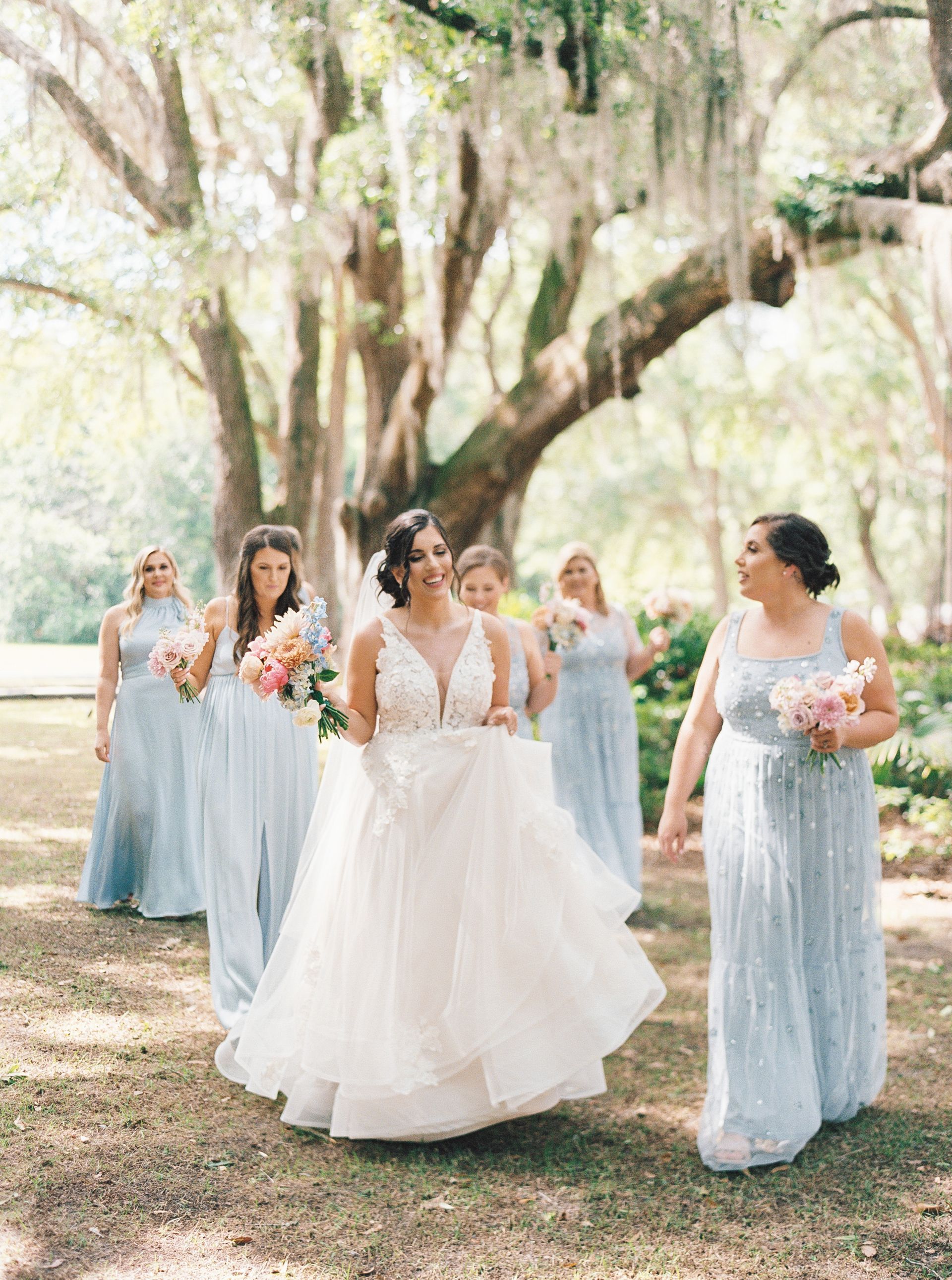 Bride with bridesmaids in light blue dresses walking outdoors under hanging moss trees.