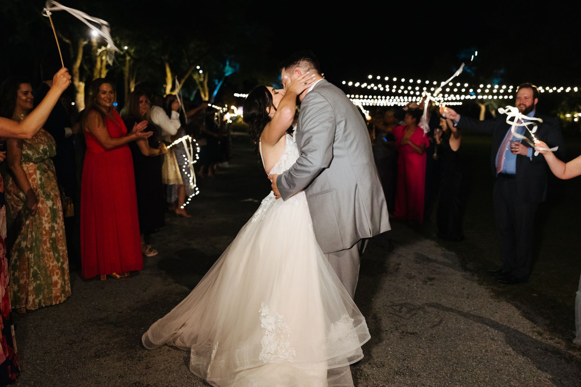 Bride and groom kissing at an outdoor nighttime wedding reception, guests cheering nearby