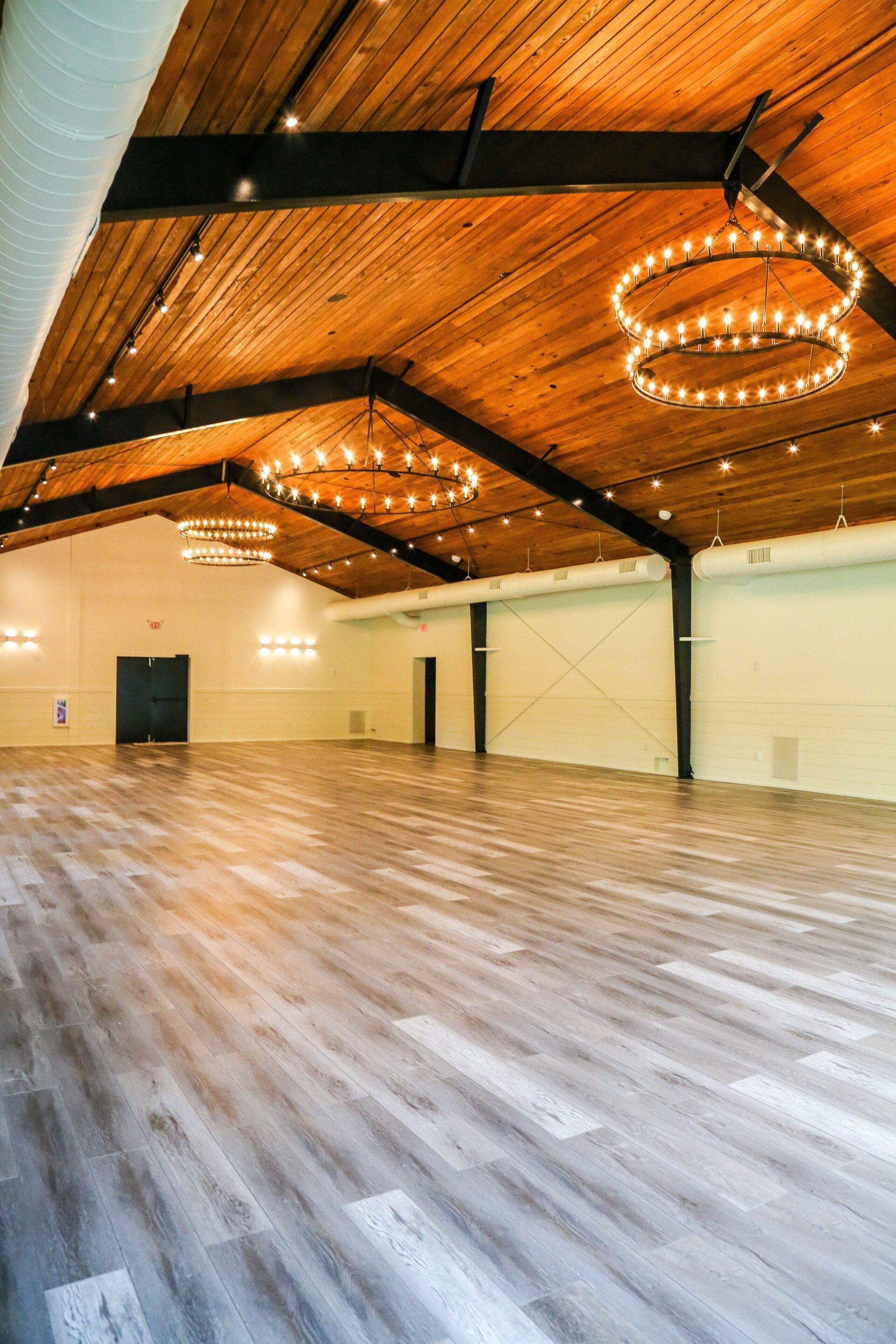 Empty rustic event hall with wood ceiling, gray floor, and two hanging ring chandeliers