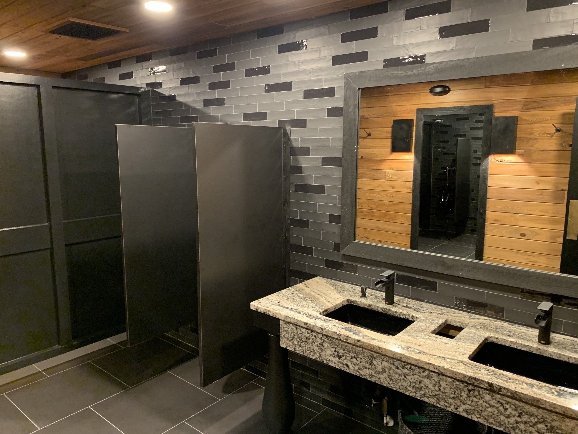 Modern restroom with dark stalls, granite double sink, and wood-and-stone wall with mirror under warm lighting