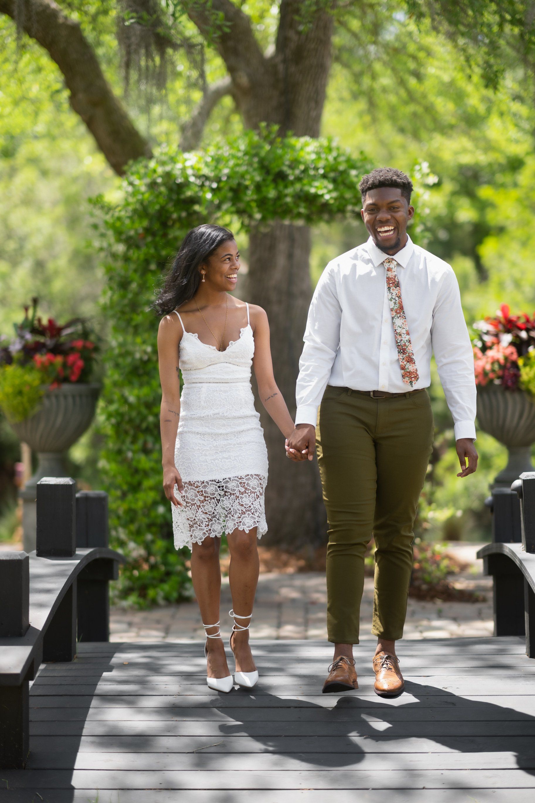 Couple holding hands on a sunlit garden bridge, dressed in white, smiling and walking together