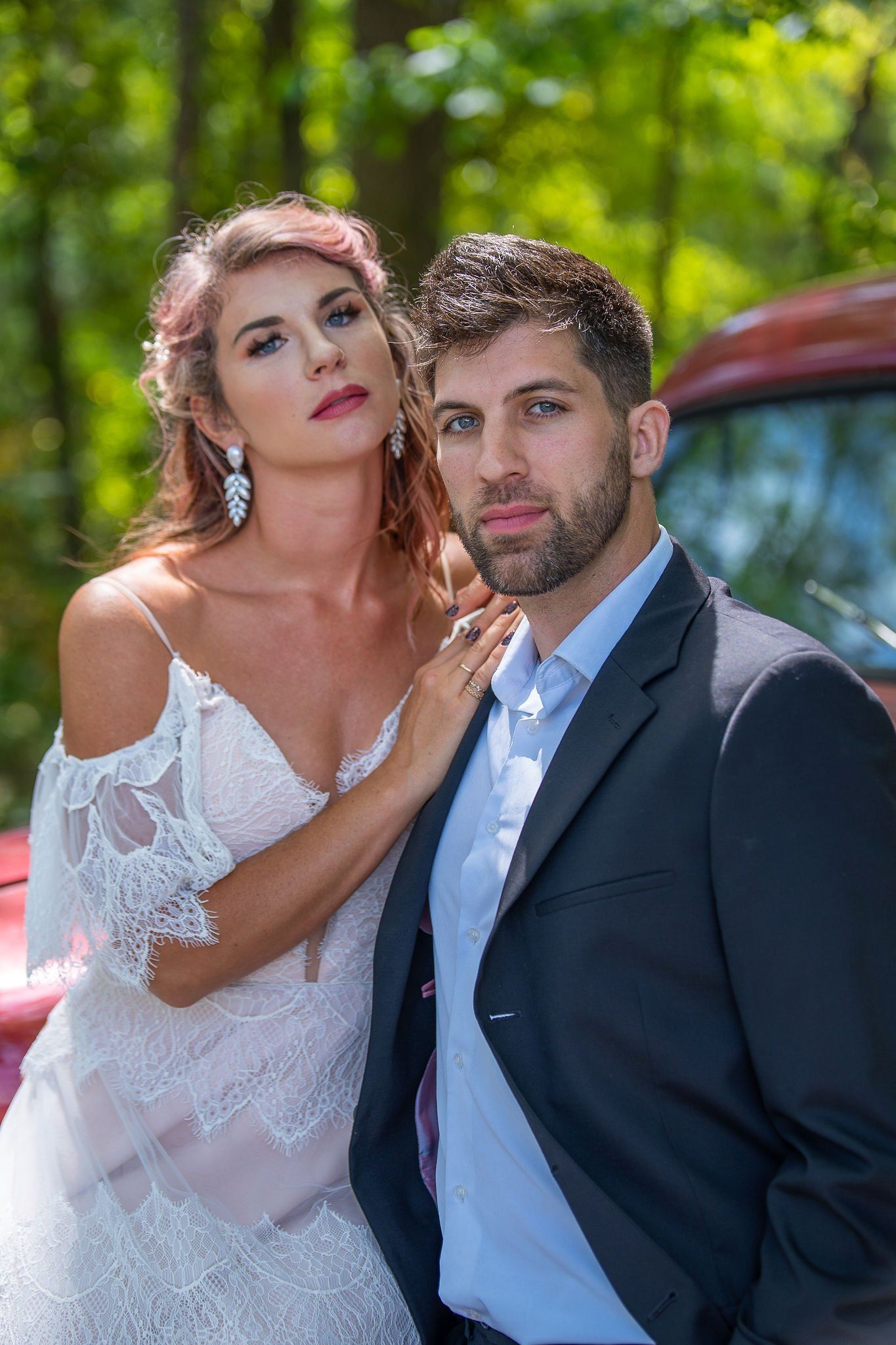 Couple posing outdoors by a red car, with a white bridal dress and dark suit in bright sunlight