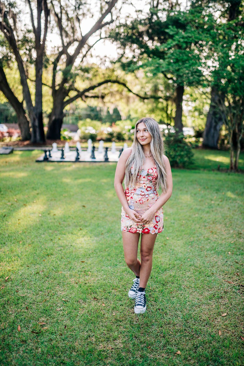 Woman in a floral dress stands on a lawn in a tree-lined park.