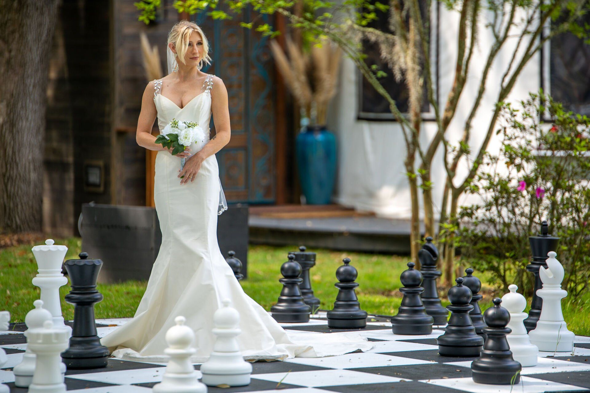 Bride in a white gown stands on a giant outdoor chessboard holding a bouquet.