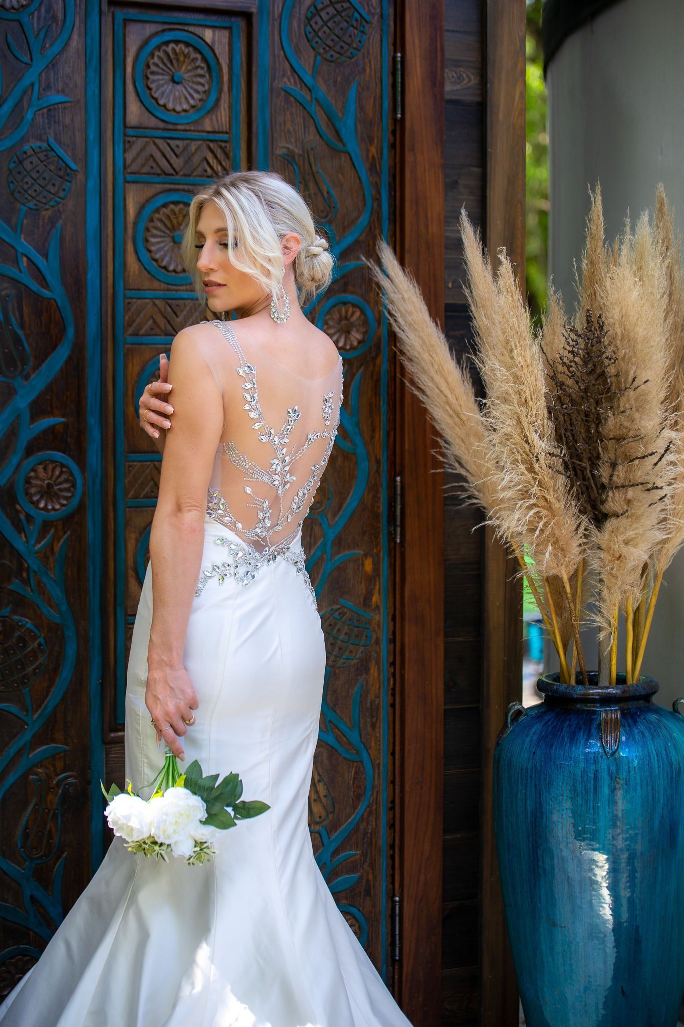 Woman in a white gown holding flowers beside an ornate wooden door and blue vase with pampas grass