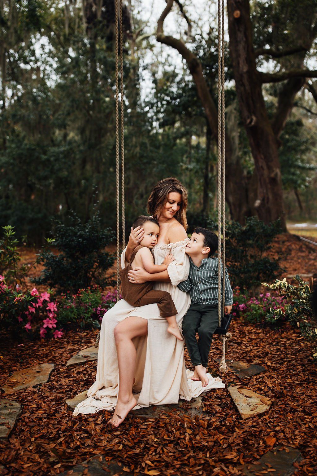 Woman seated in a wooded garden with two children, surrounded by flowers and fallen leaves.