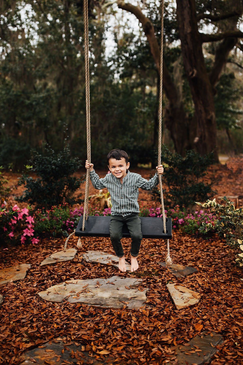 Child smiling on a swing in a wooded garden, surrounded by fallen leaves and pink flowers.