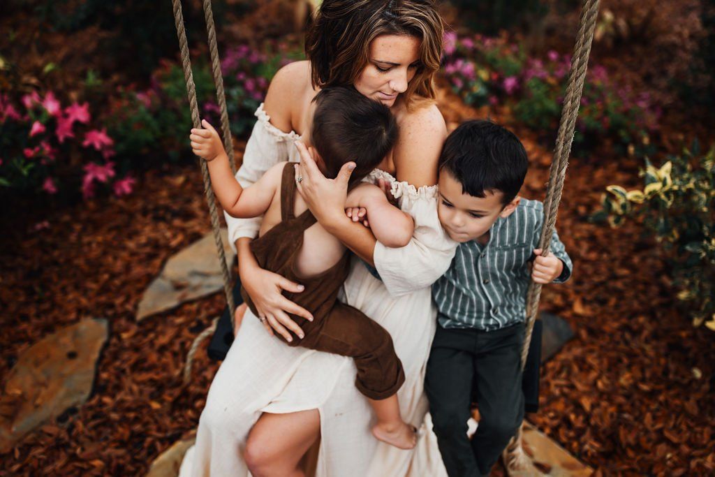 Woman on a swing holding two children in a leafy outdoor setting.