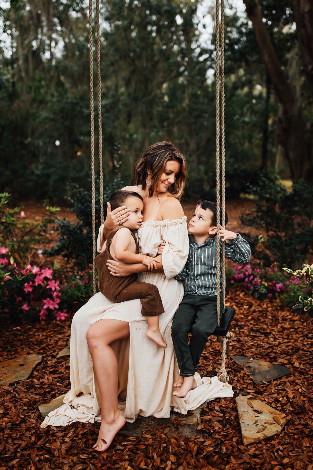 Woman sitting on a swing holding two children in a flower-filled garden with tall trees