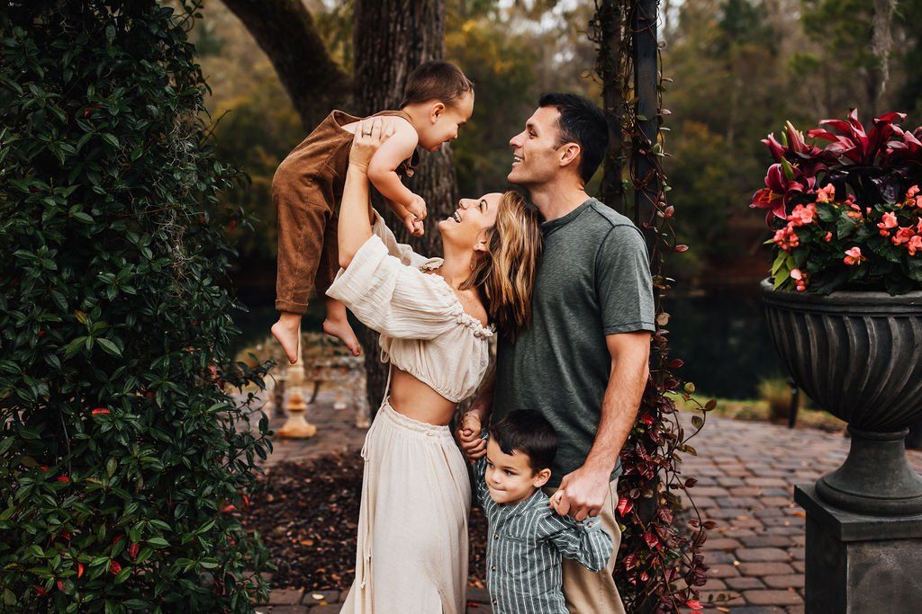 Family posing in a garden path, with parents lifting two children near lush greenery and flowers.