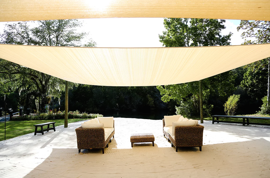 Outdoor lounge area with wicker sofas and a beige canopy on a sandy patio, surrounded by trees