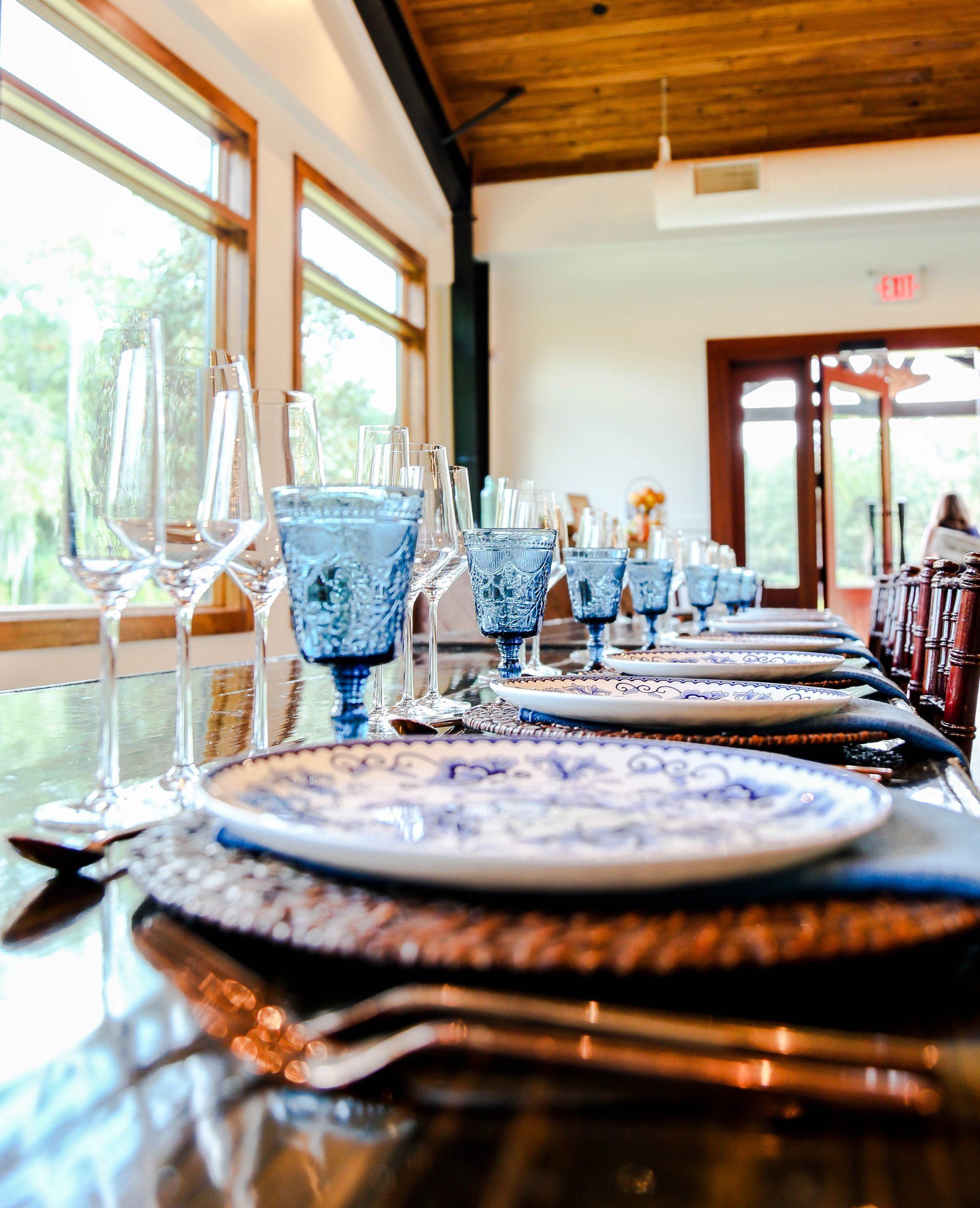 Elegant dining table set with blue glassware, patterned plates, and silverware by large windows in a bright room