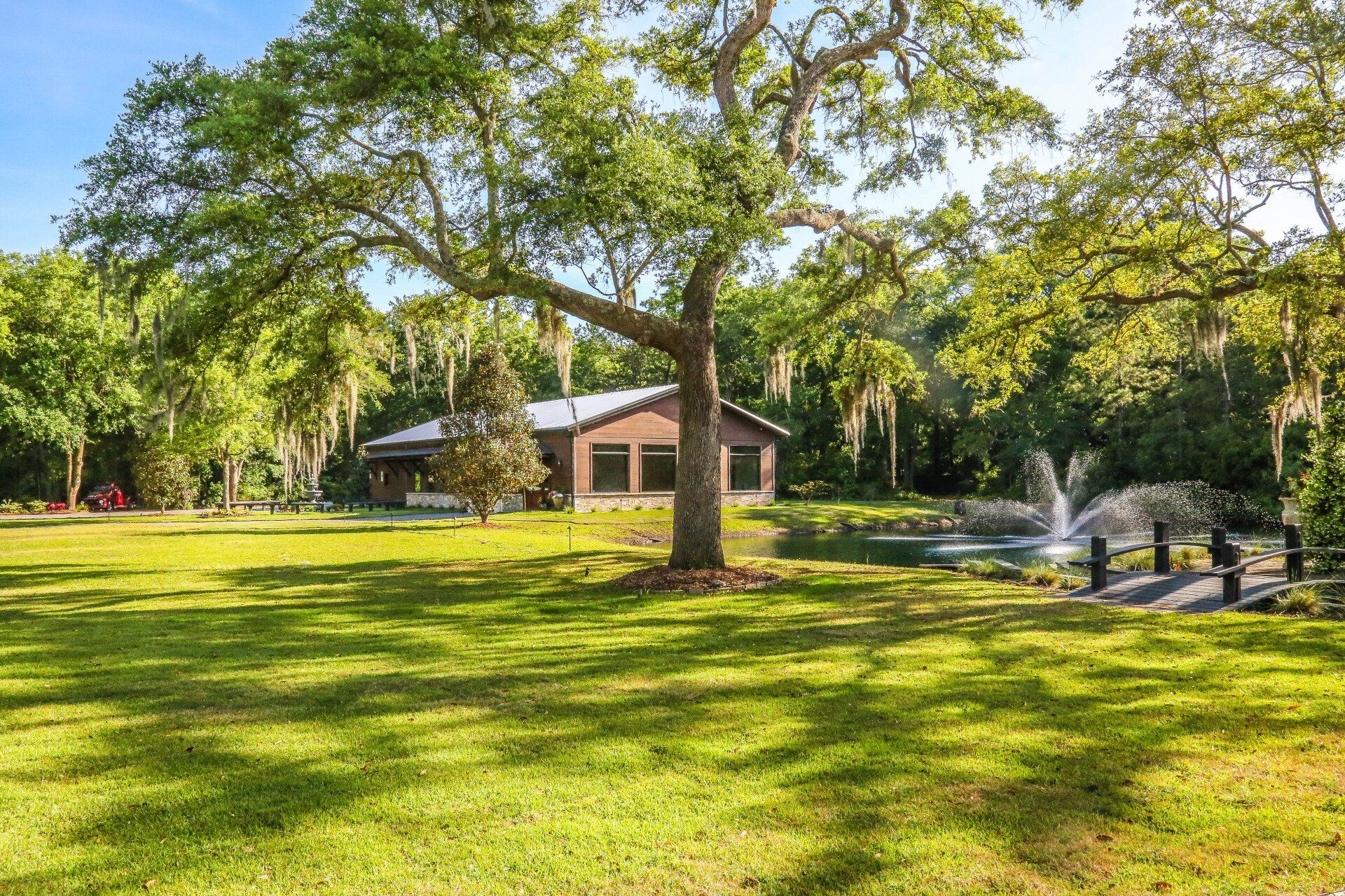 Sunny park with green lawn, large trees, a small building, and a fountain spraying water