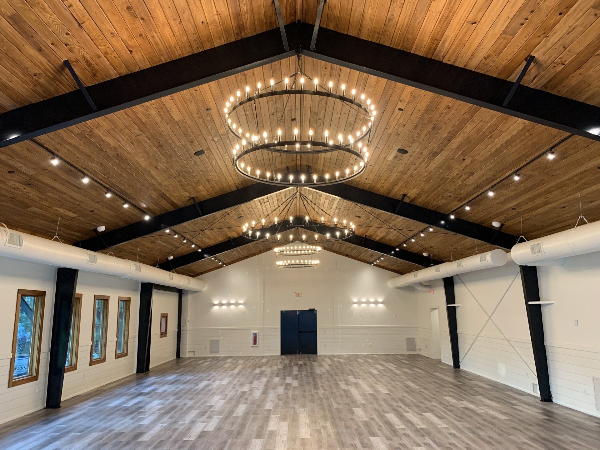 Spacious empty hall with wood-beamed ceiling, chandeliers, white walls, and wood flooring