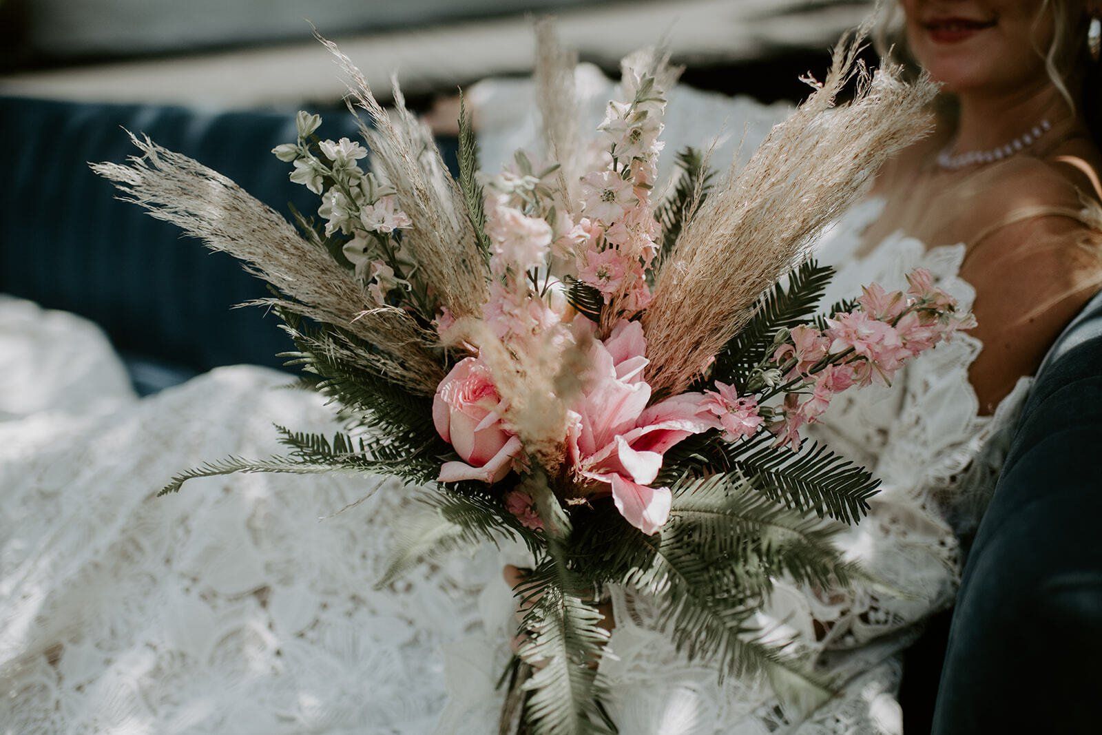 Bride holding a pastel pink and beige bouquet with pampas grass, seated in a white lace dress