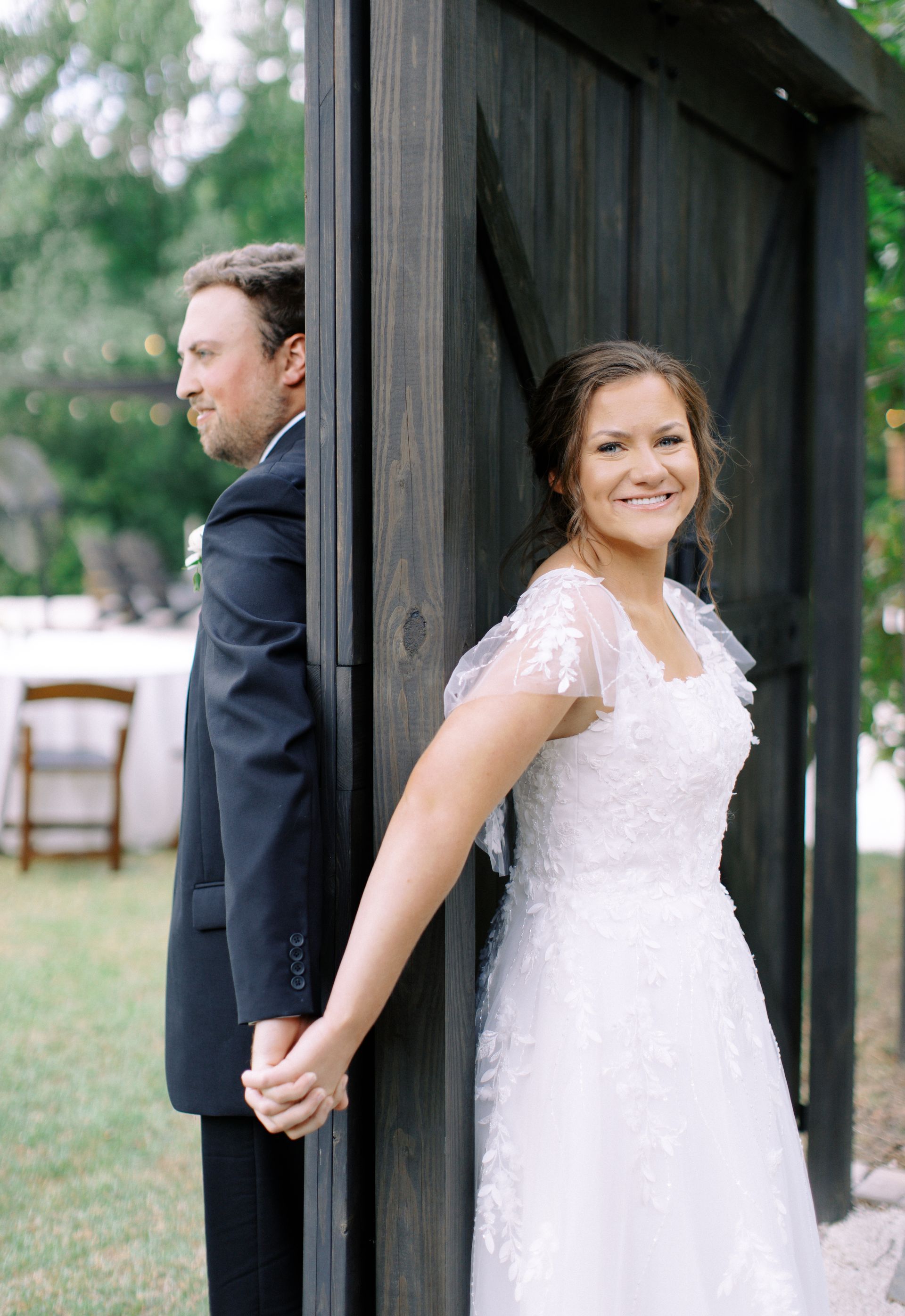 Bride and groom stand back-to-back by a wooden structure, holding hands and smiling outdoors.